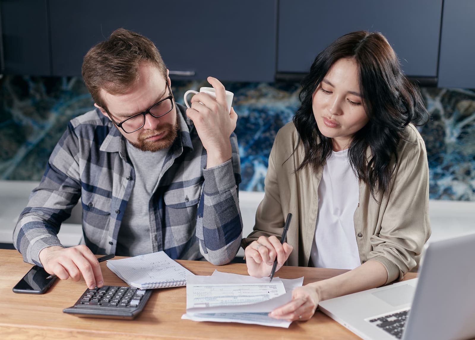 A couple at a kitchen table working through paperwork with a calculator, a notebook and a laptop