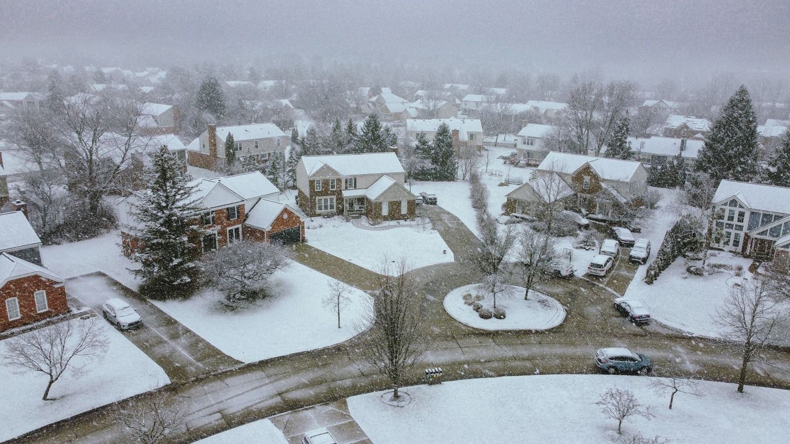 A suburban cul-de-sac of brick houses under fresh snow, seen from above