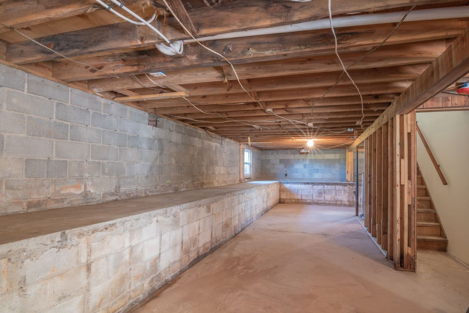An unfinished basement with concrete block walls, exposed wooden joists, wiring along the ceiling and a wooden staircase