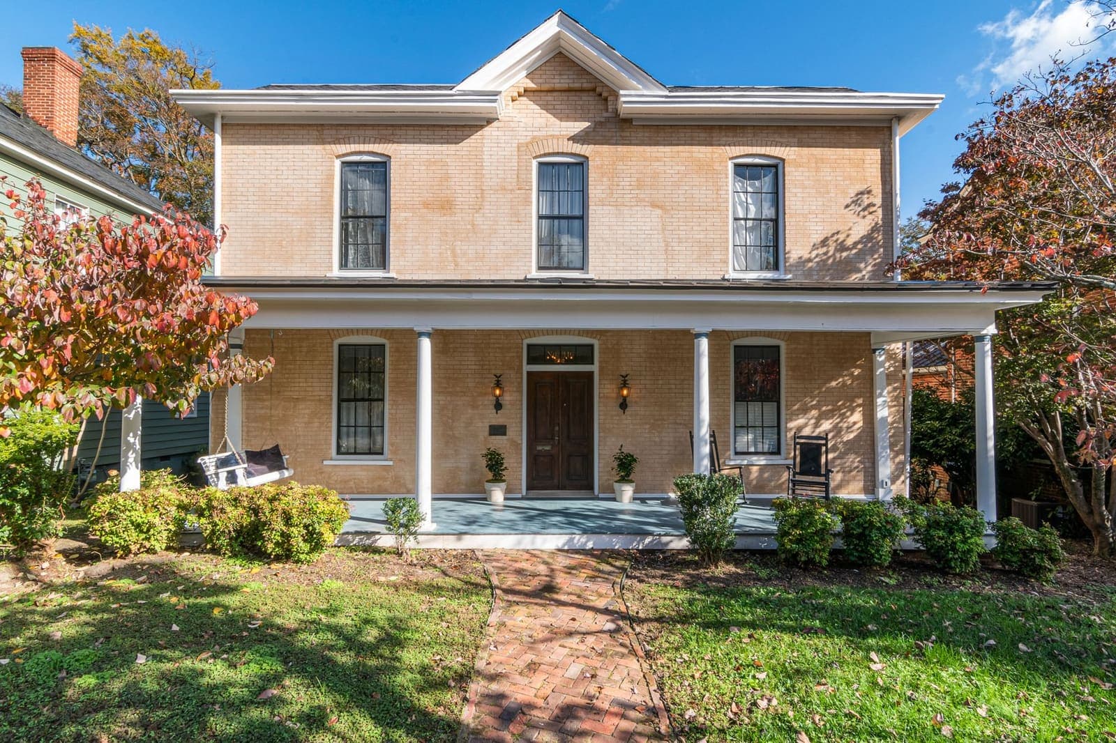 A two-storey brick house with a deep front porch, a porch swing and rocking chairs, with autumn trees on either side