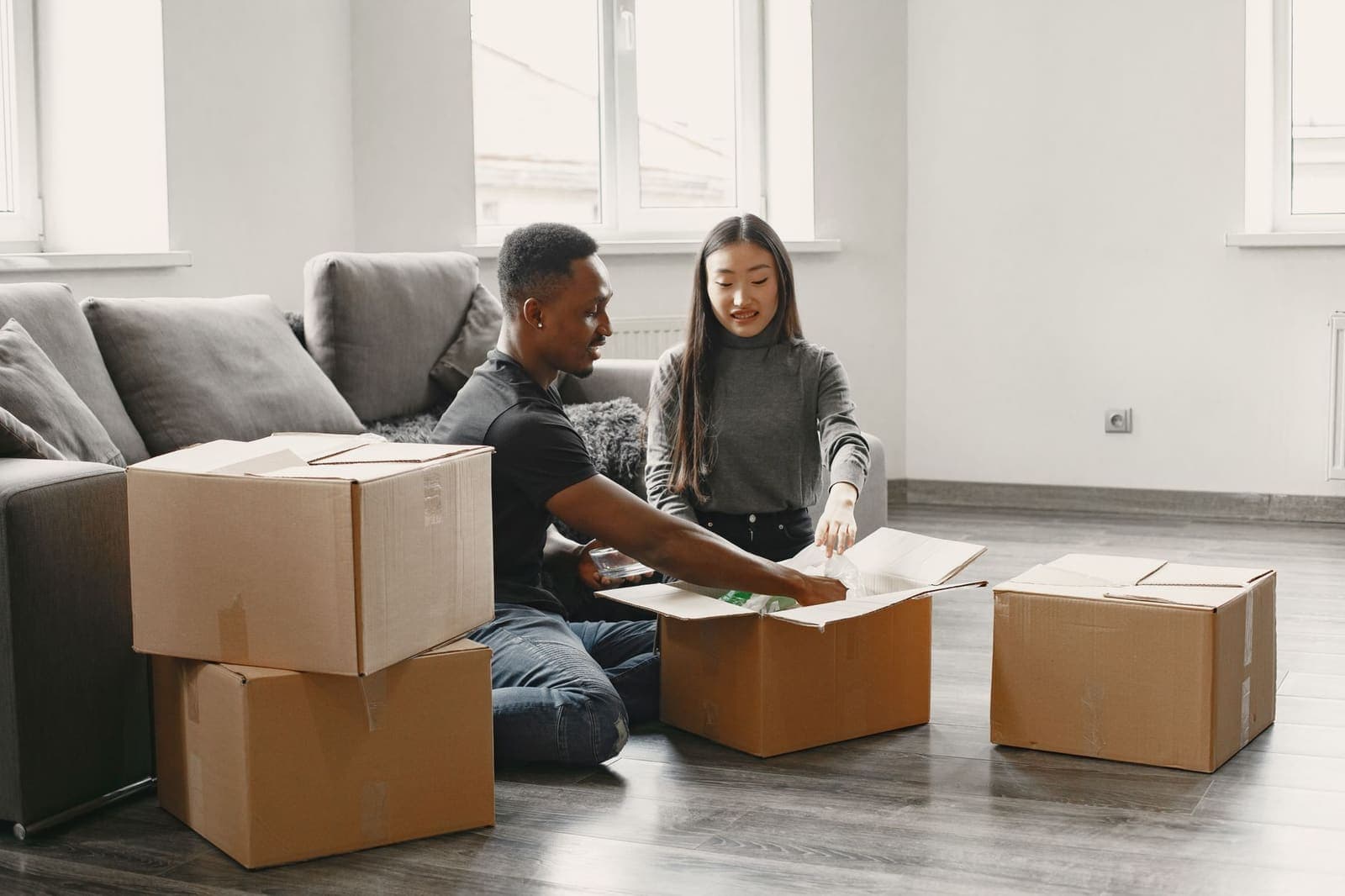 Two people sit on a living room floor unpacking cardboard moving boxes beside a sofa