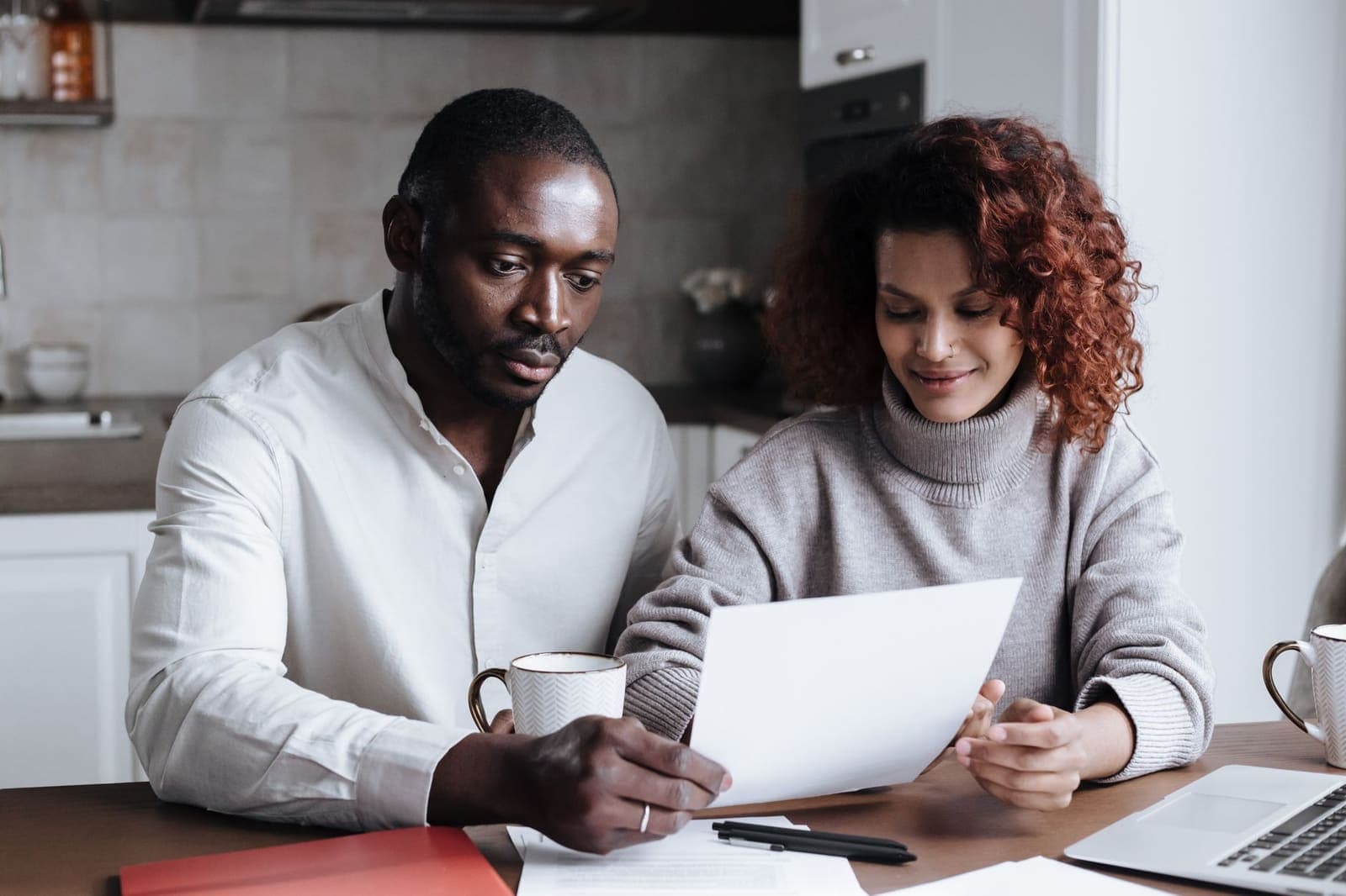 Two people at a kitchen table read a printed document together, with a laptop and papers beside them