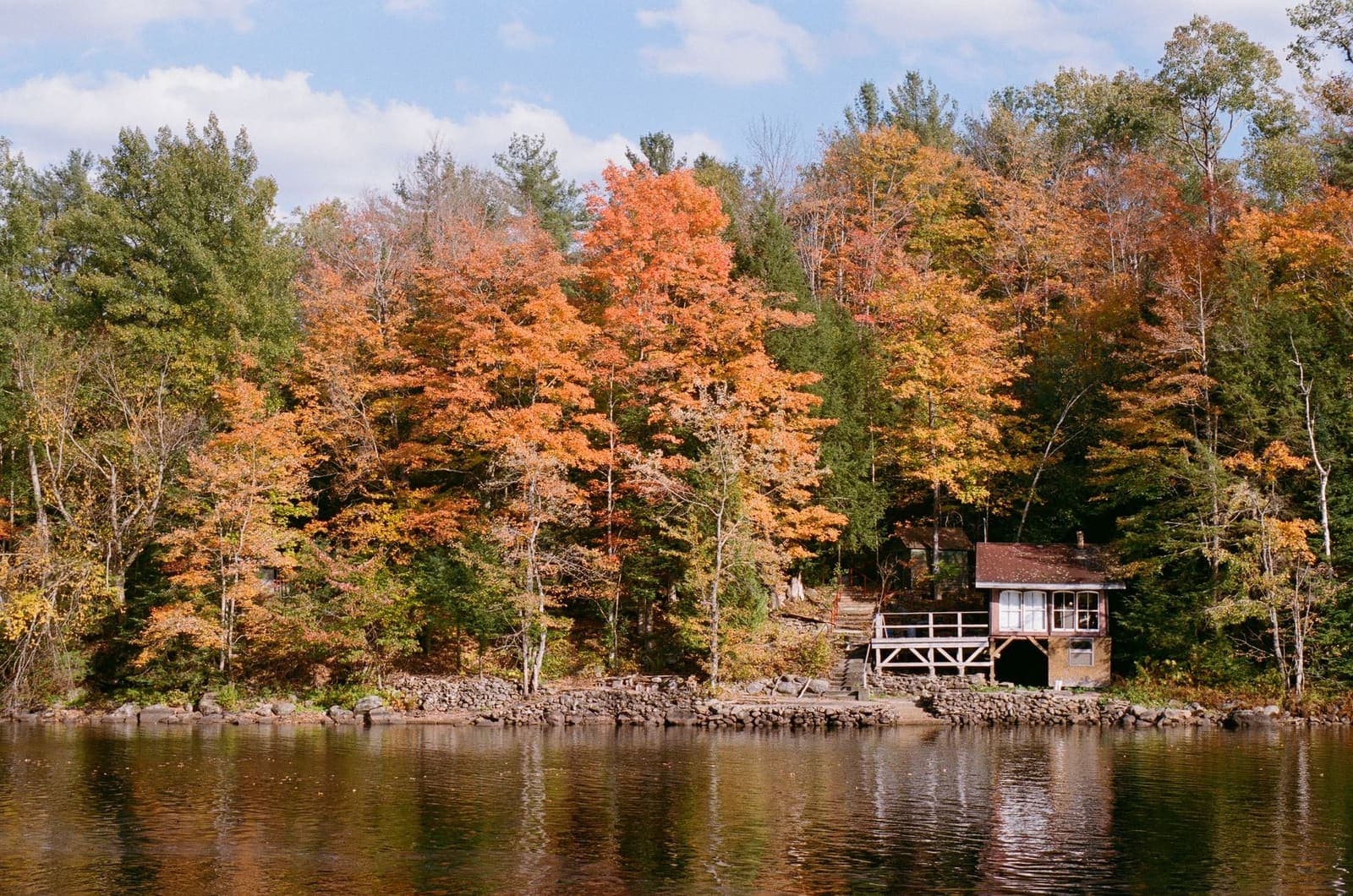 A small wooden cabin with a deck on a lakeshore, set against a hillside of orange and yellow autumn trees