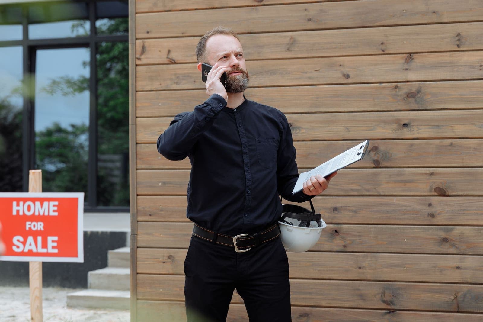 A man in a dark shirt holds a phone to his ear and a clipboard outside a house with a Home for Sale sign