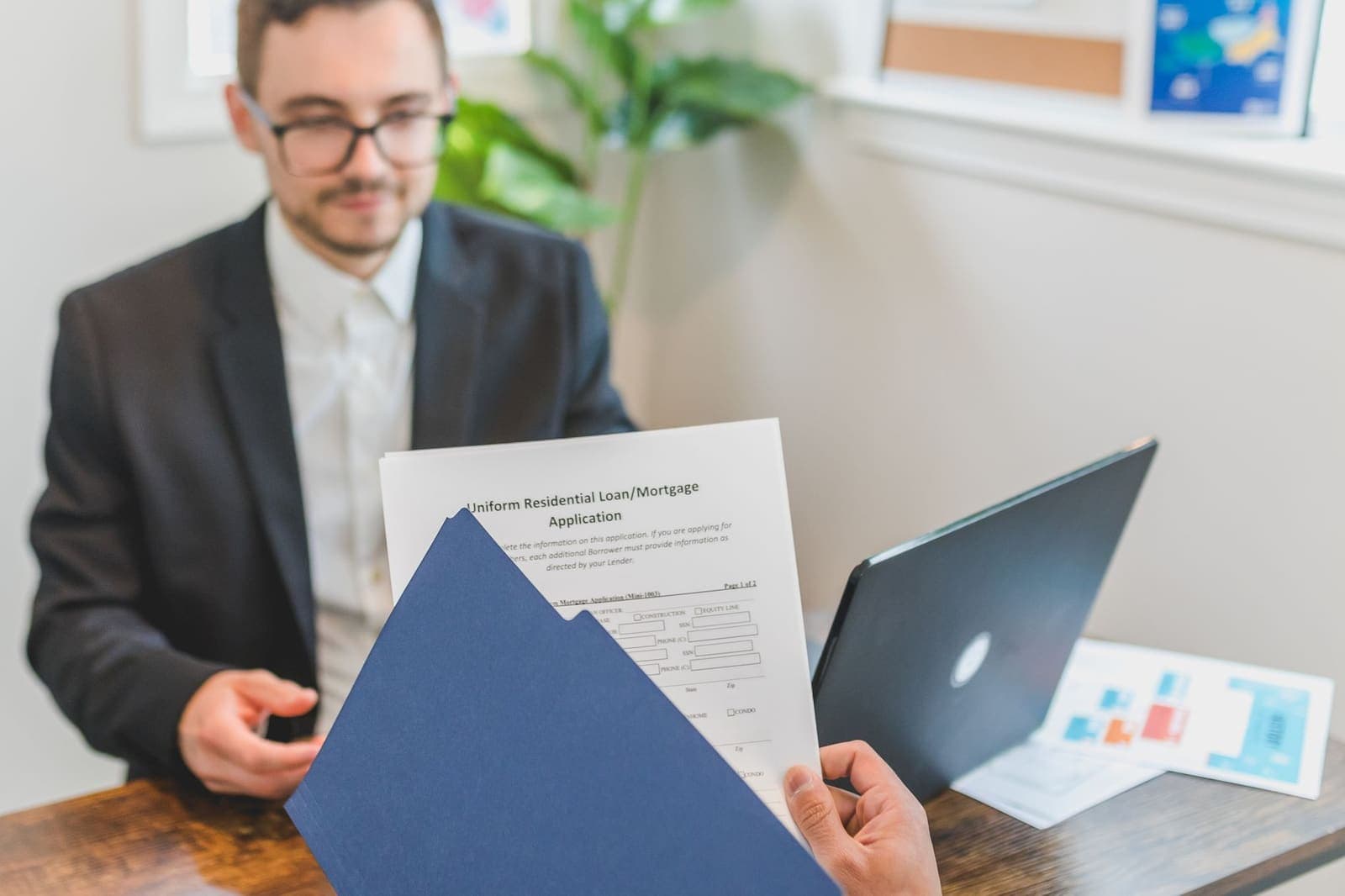 A loan officer at a desk across from a client holding a Uniform Residential Loan Application in a blue folder