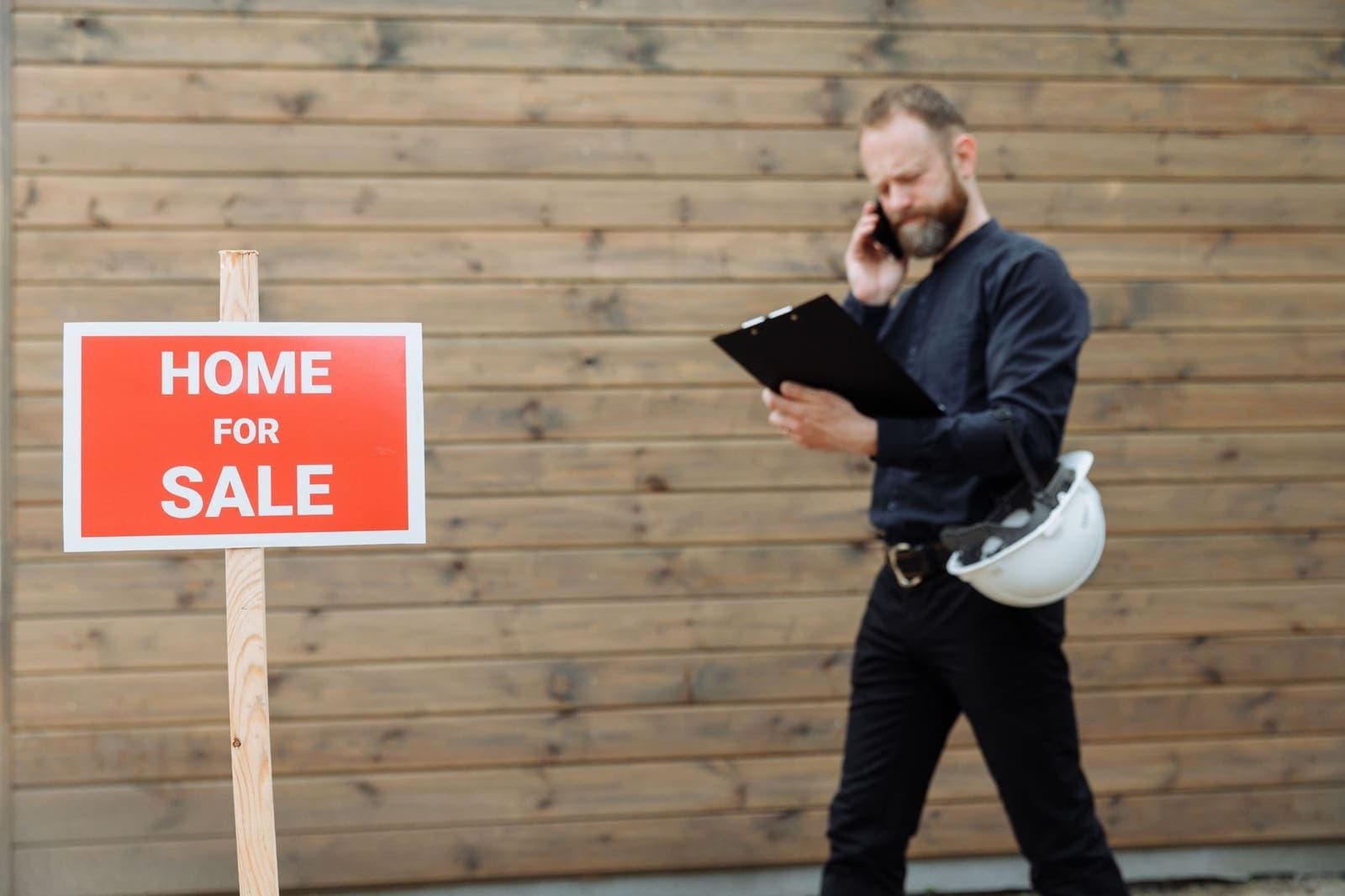 A real estate agent on the phone reads a clipboard beside a red Home for Sale sign on a wooden stake