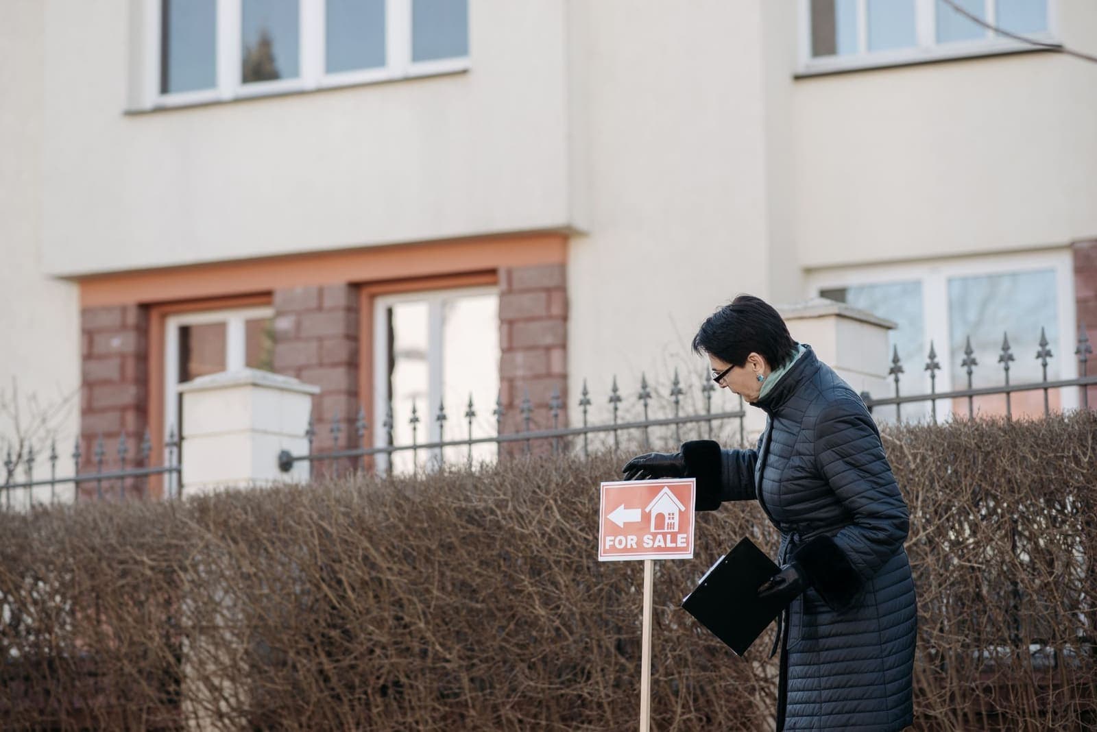 A real estate agent in a dark winter coat adjusts a small For Sale sign in front of a hedge and a two-storey house