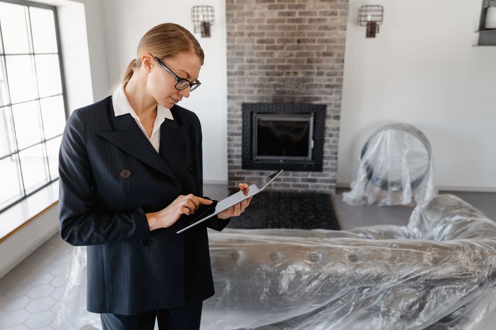 A real estate agent in a pinstripe suit and glasses works on a tablet in an empty living room with furniture under plastic sheeting