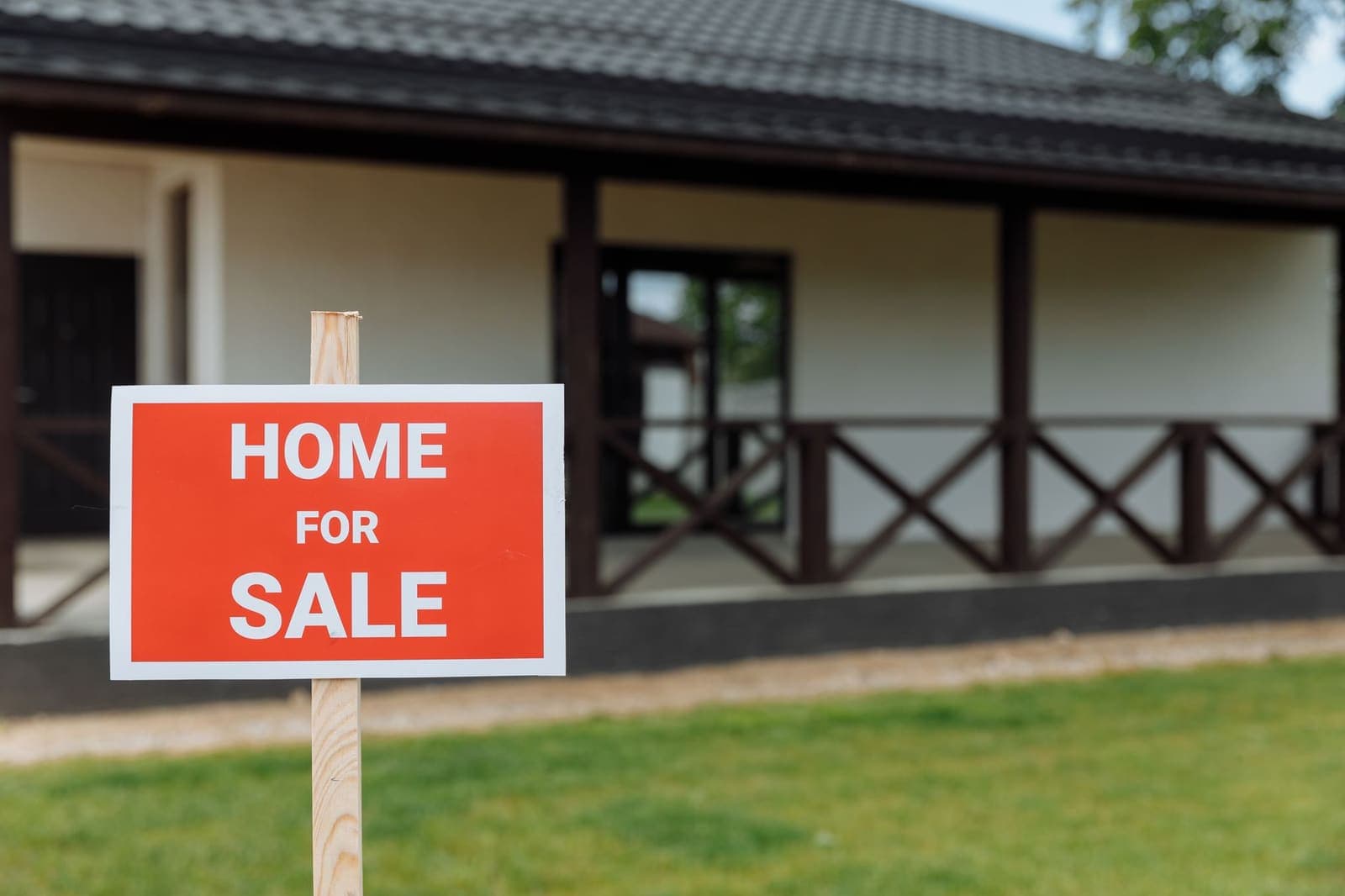 A red Home For Sale sign on a wooden stake in the front lawn of a single-storey house with a covered porch