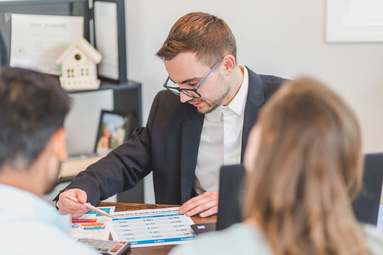 A loan officer in a dark jacket pointing with a pen at a printed rate table while a couple sits across the desk