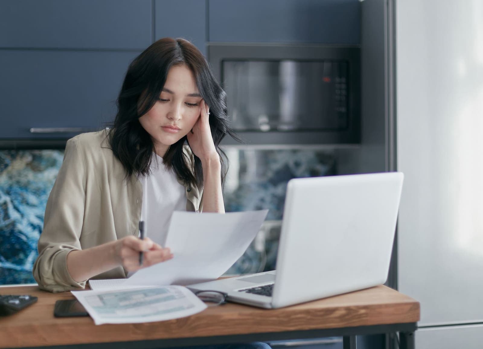 A woman at a kitchen table reading a printed form beside a laptop with more paperwork in front of her