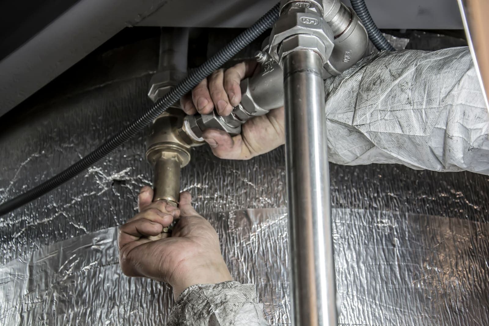 A plumber's hands tightening a brass fitting on steel pipework under a sink