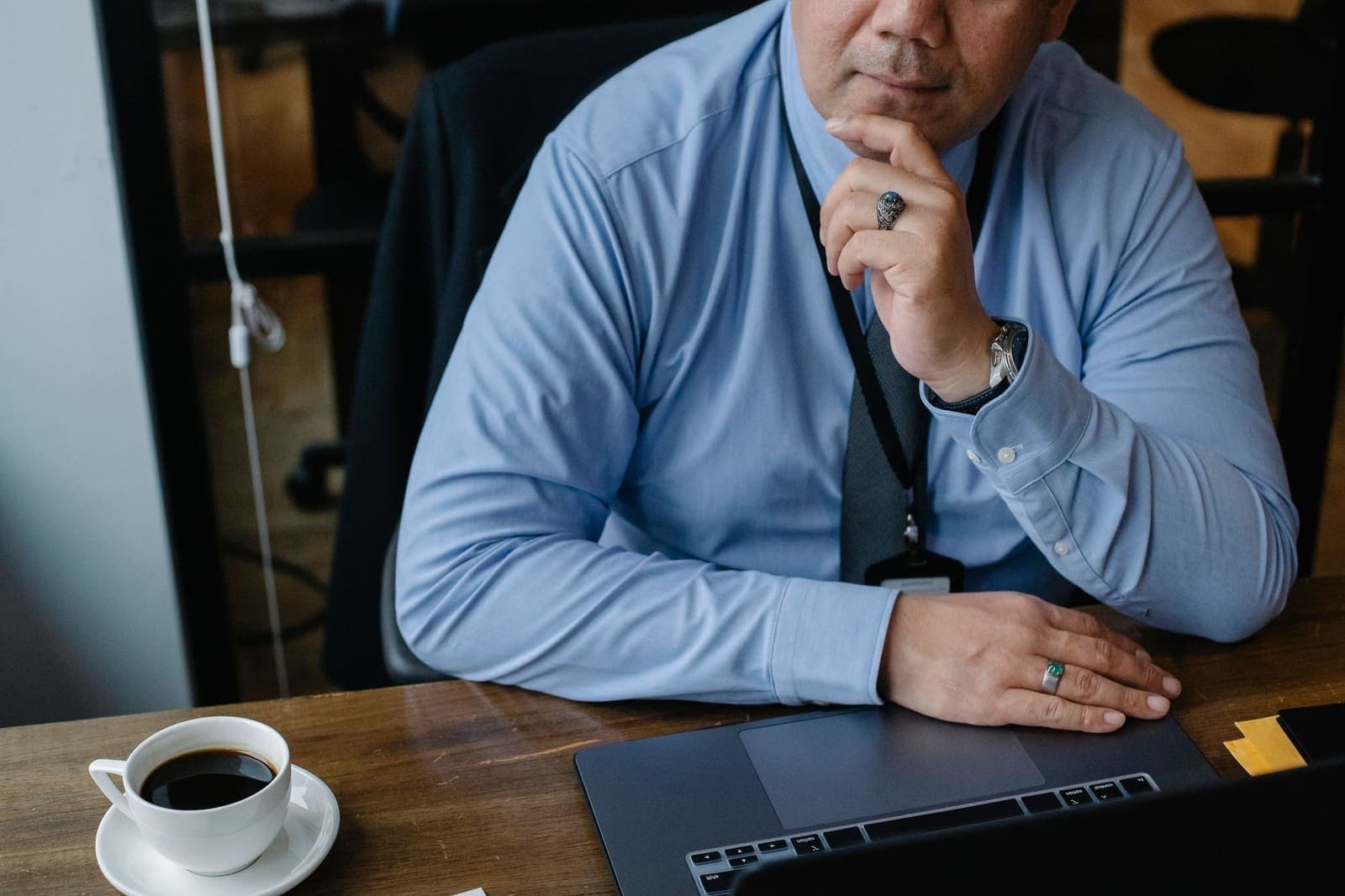 A man in a blue shirt sits at a wooden desk with a laptop and a cup of coffee, hand on his chin