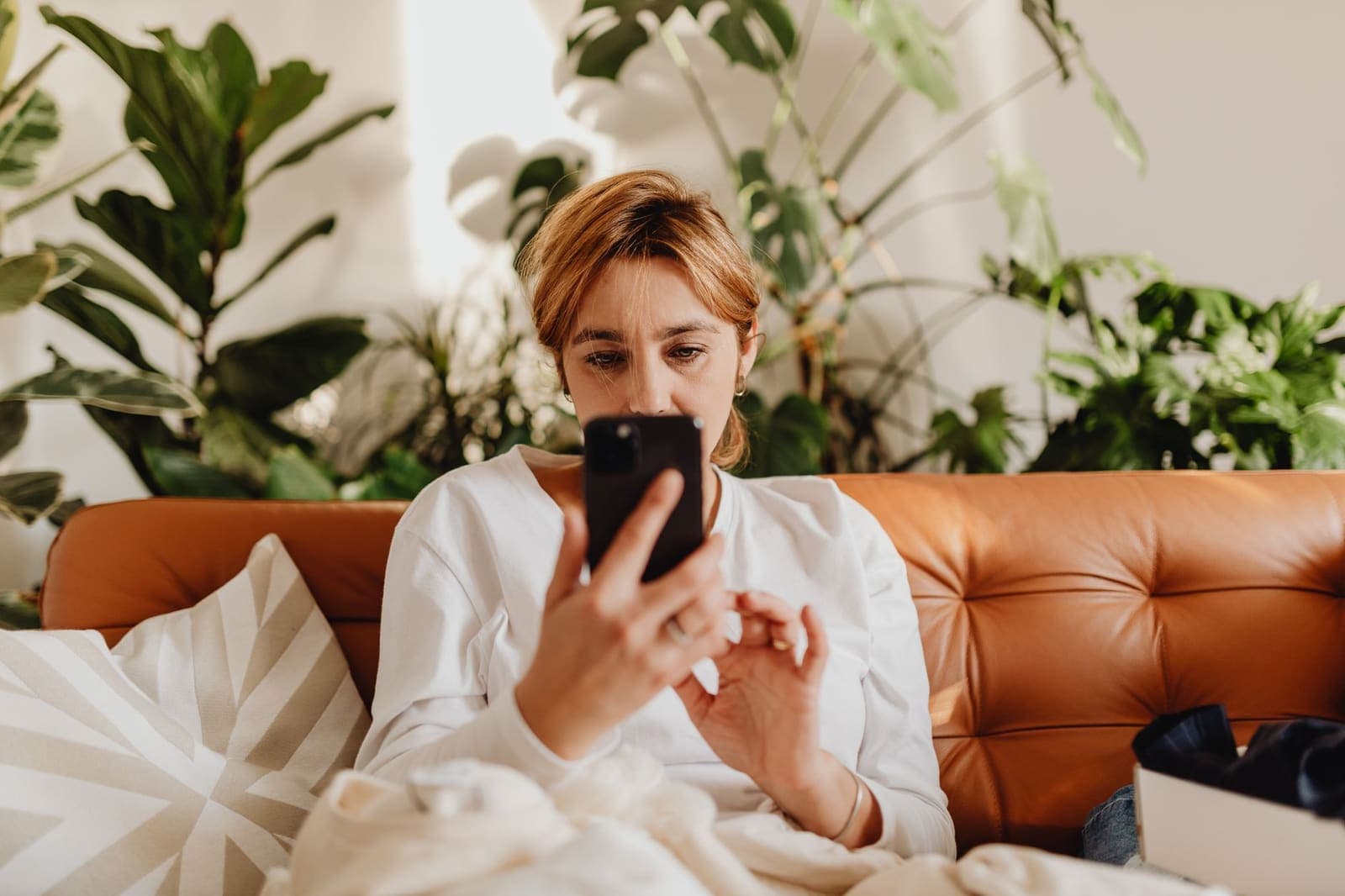 A woman on a leather sofa looks closely at her phone, with house plants behind her