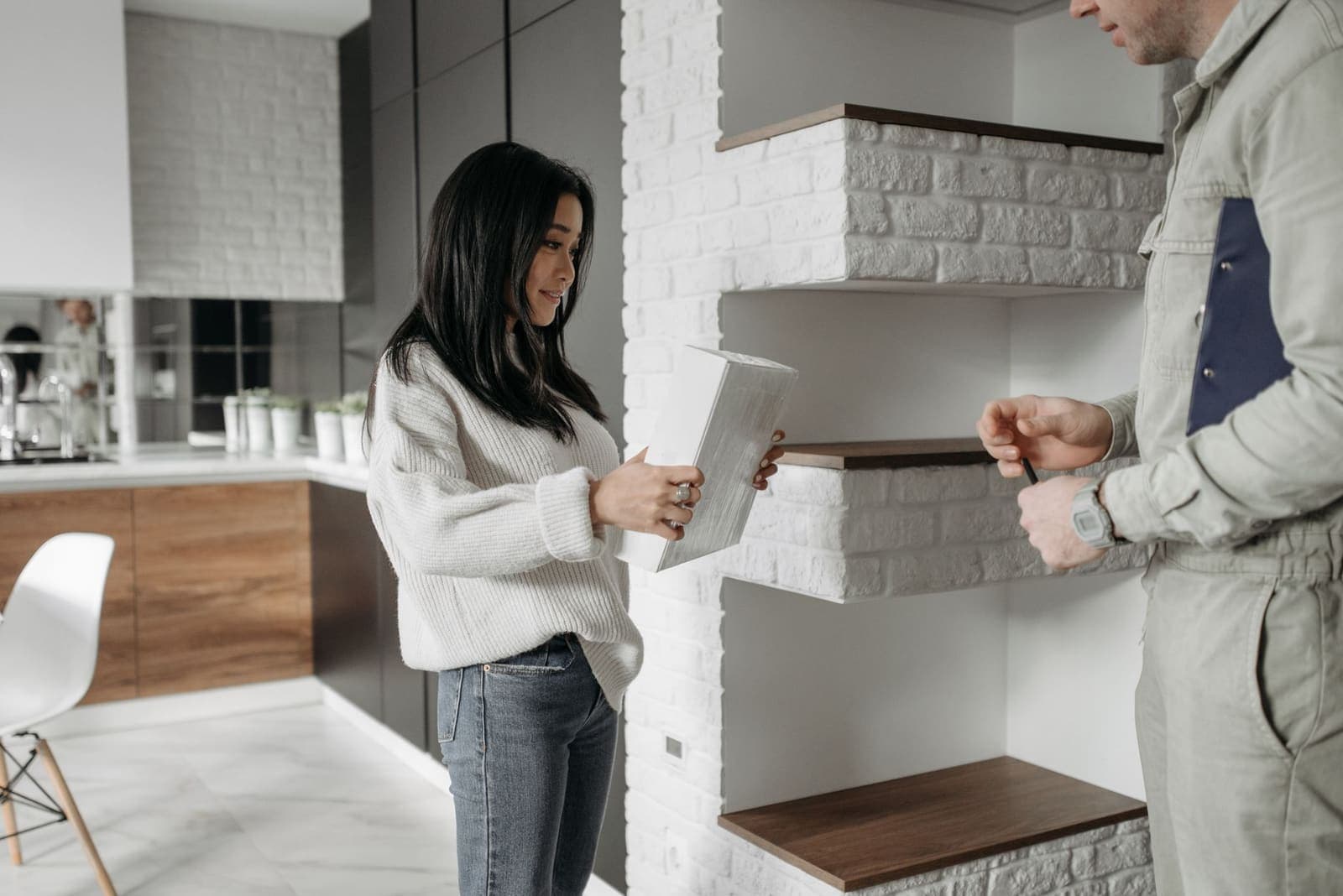 A tradesman in grey overalls with a clipboard talks with a homeowner who holds a small box beside open wall shelving