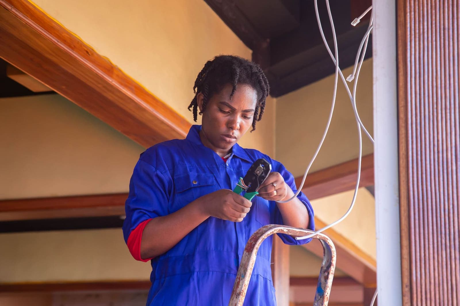 A technician in blue overalls stands on a stepladder and crimps the end of a white cable