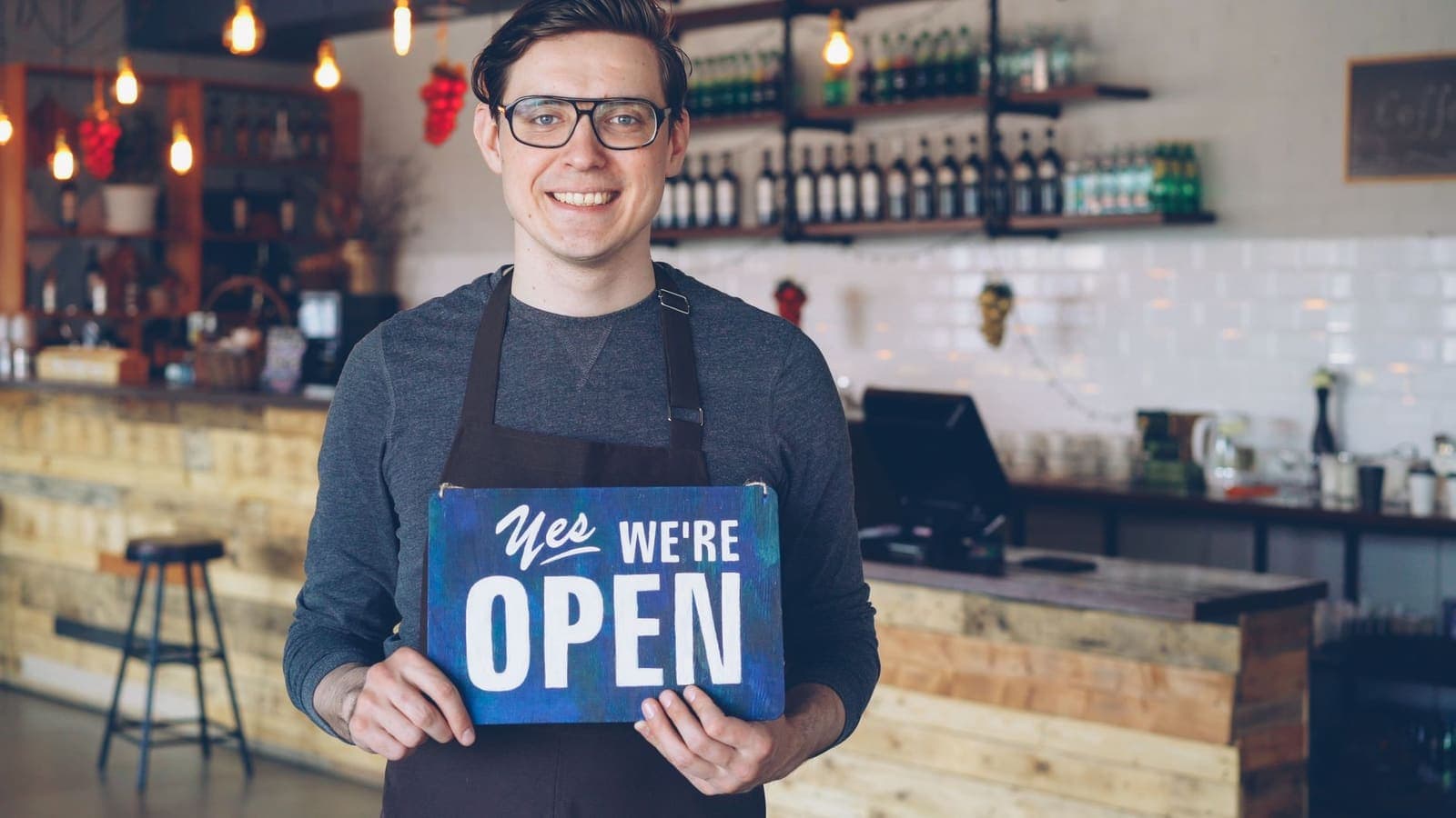 A café owner in an apron holds up a hand-painted sign reading yes we're open in front of the counter
