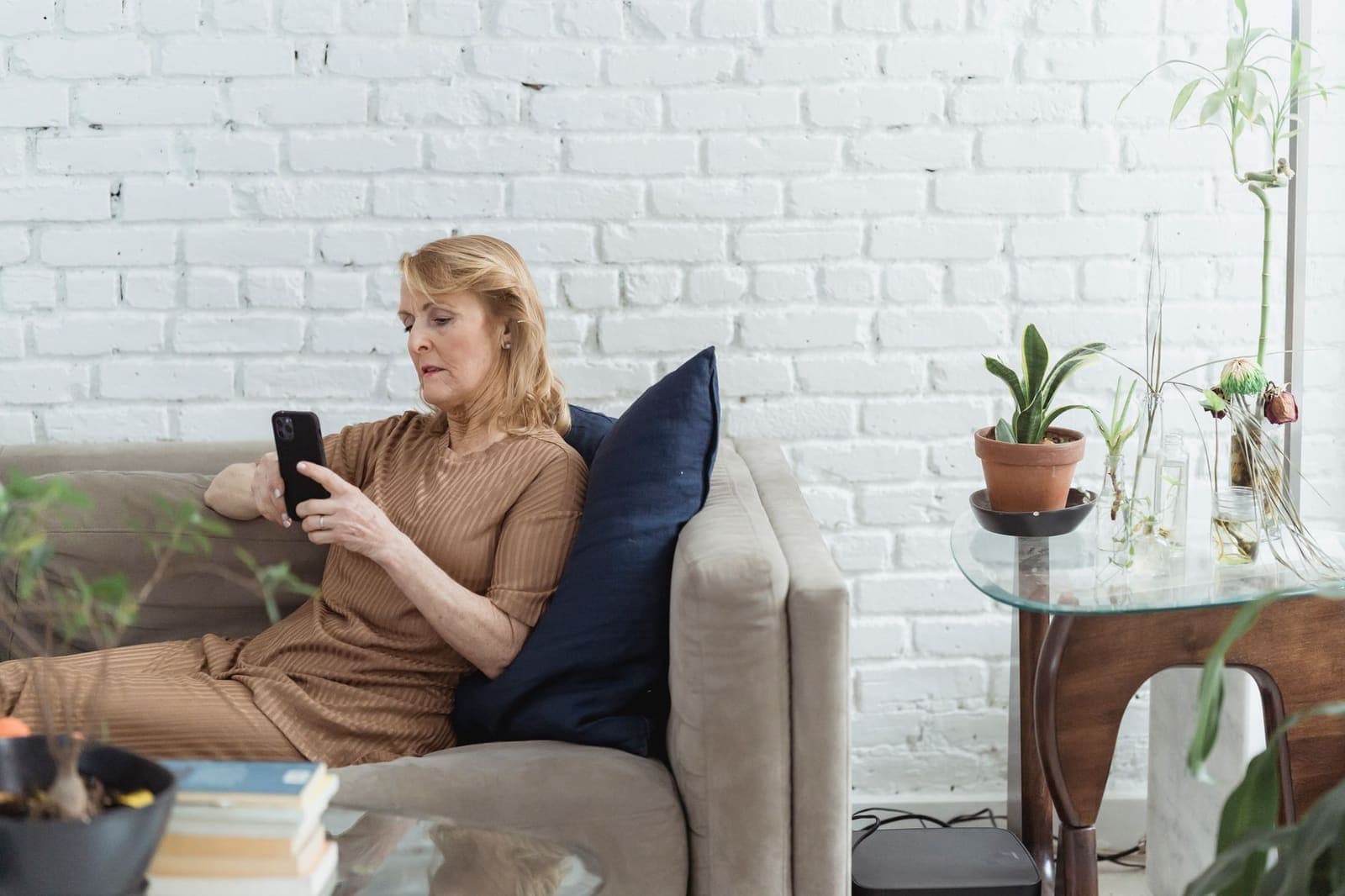 An older woman sitting on a sofa by a white brick wall, looking at her phone