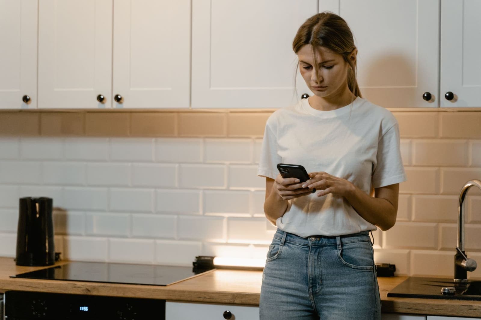 A woman in a white T-shirt stands at a kitchen counter in the evening, looking at her phone