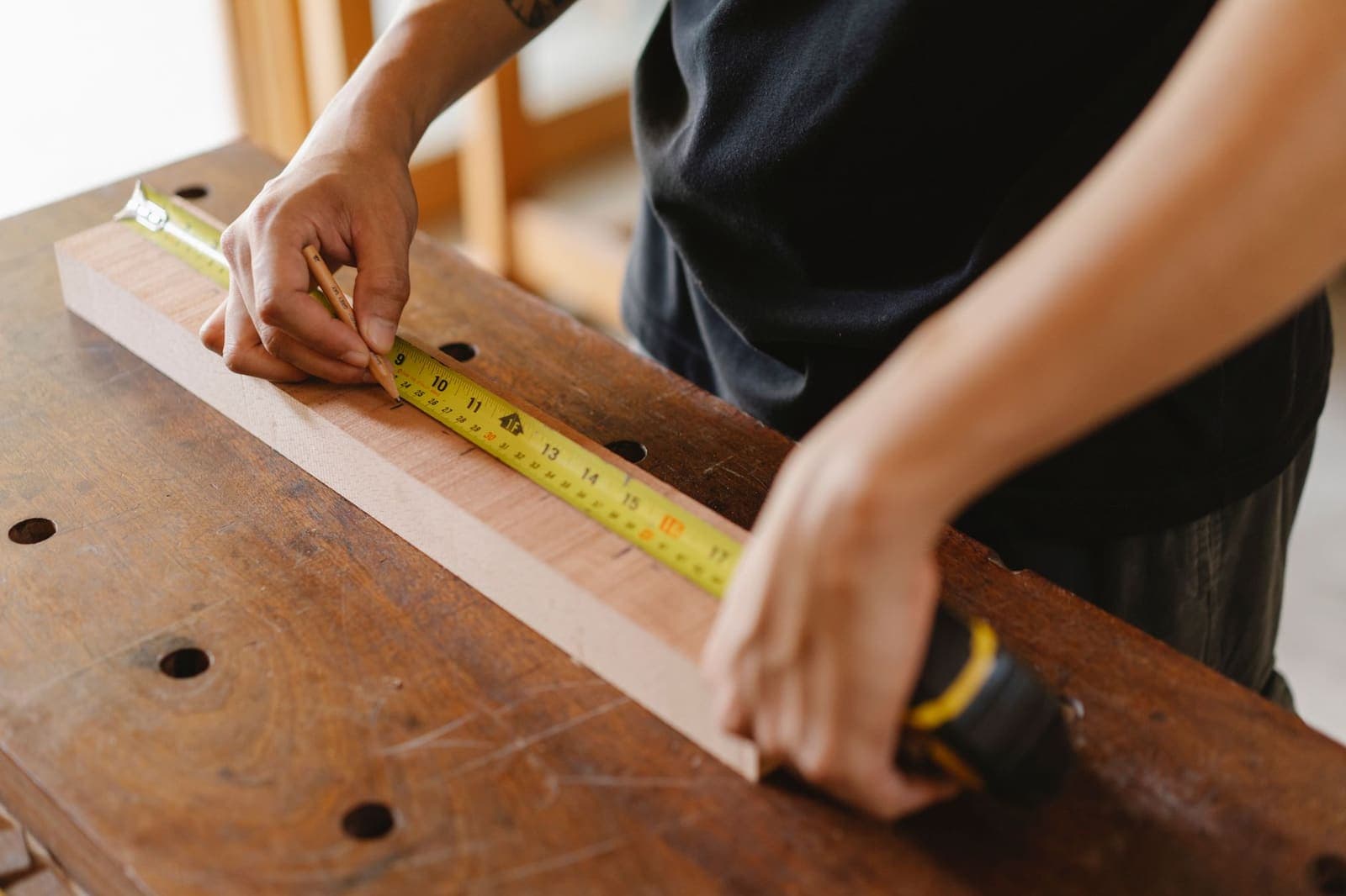 A carpenter marks a wooden board with a pencil against a yellow tape measure on a workbench
