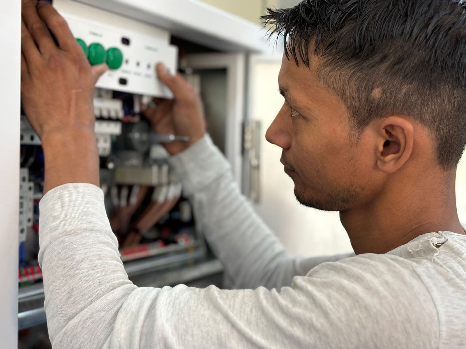 An electrician in a grey long-sleeved shirt works on the breakers inside an open electrical panel