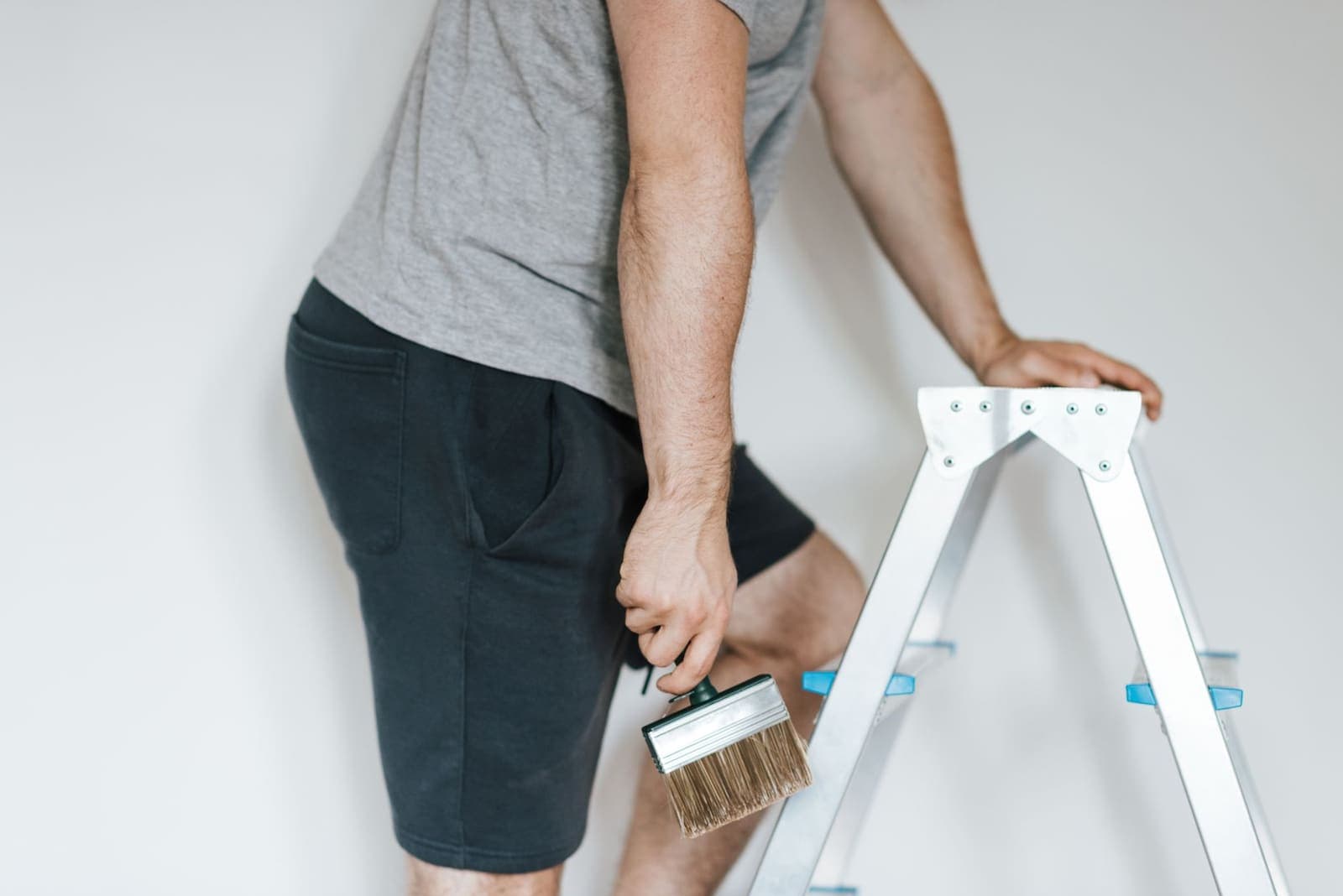 A painter in a grey T-shirt holds a wide brush and steadies himself on an aluminium stepladder against a white wall