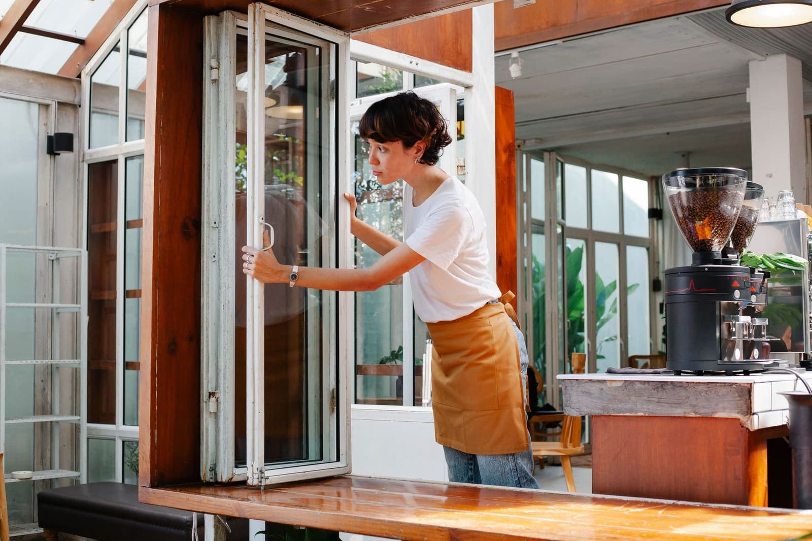 A young worker in an apron opening the folding window of a cafe at the start of a shift