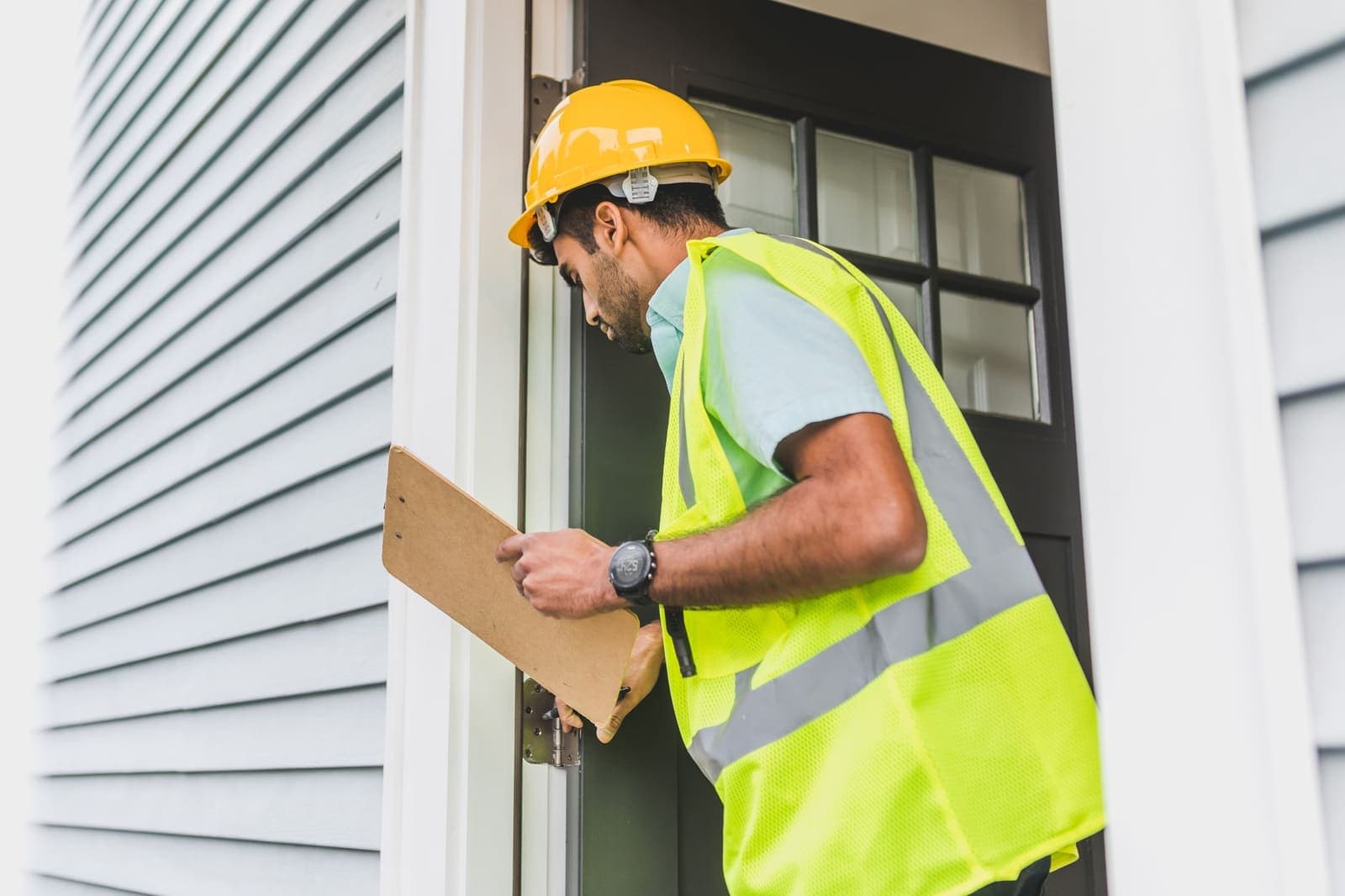 A building inspector in a yellow hard hat and high-visibility vest checking a doorway with a clipboard
