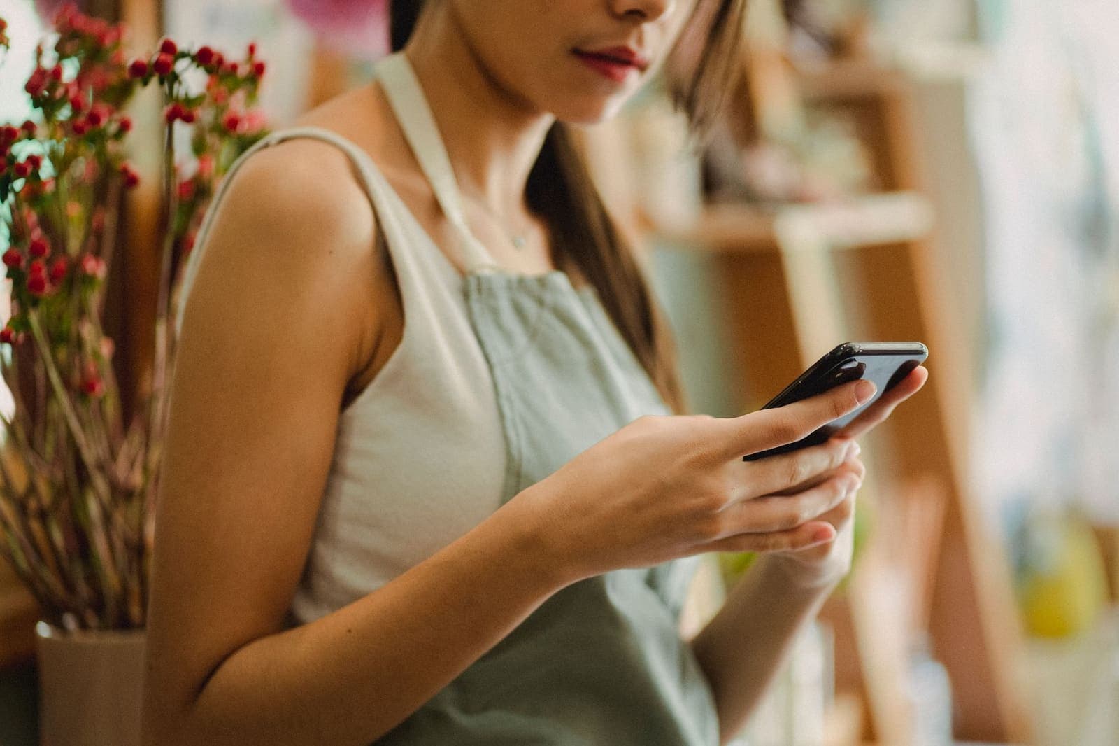 A florist in an apron reads a message on a smartphone beside a vase of red berries