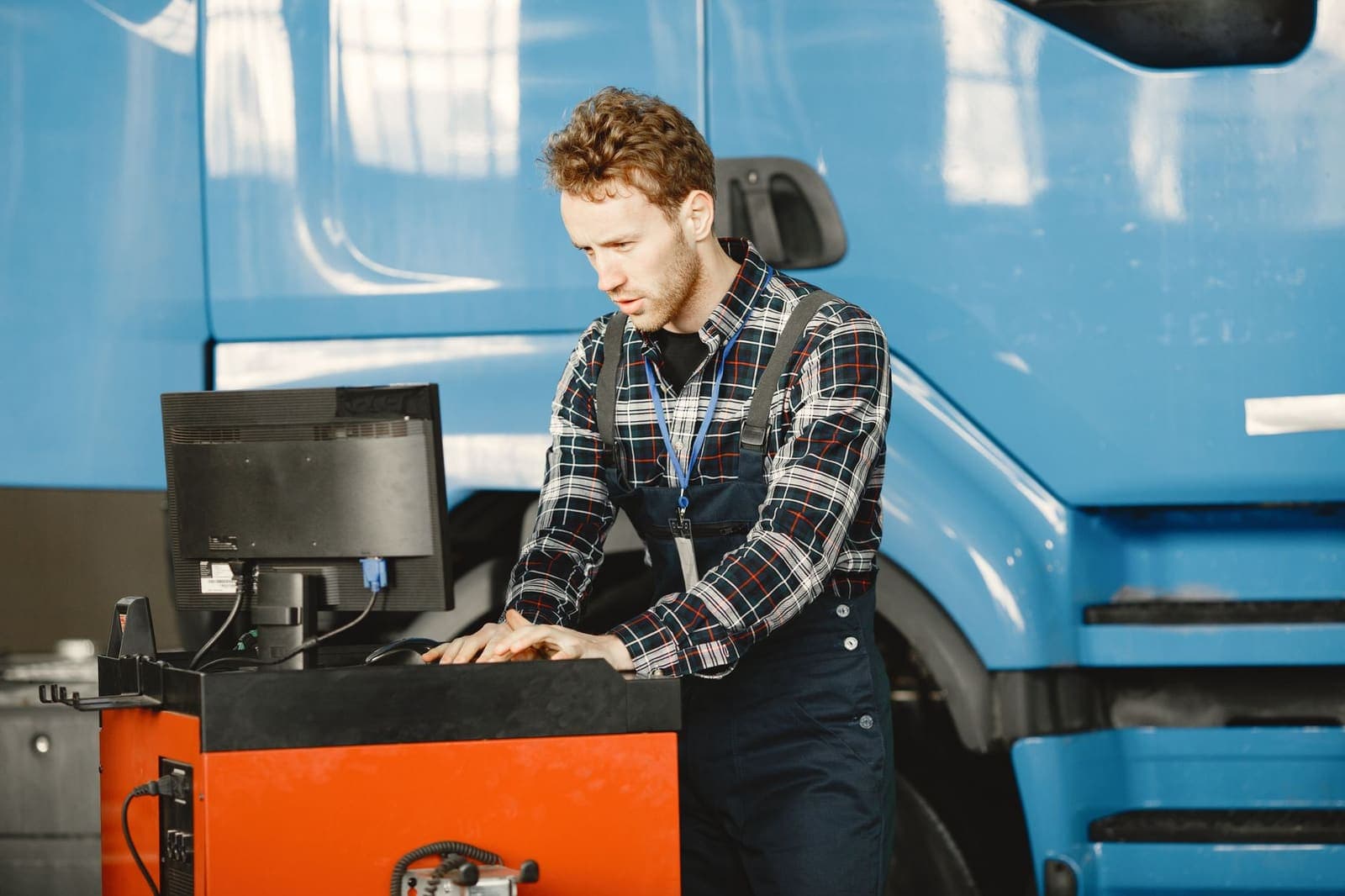 A mechanic in overalls types at a workshop computer station beside a blue truck cab