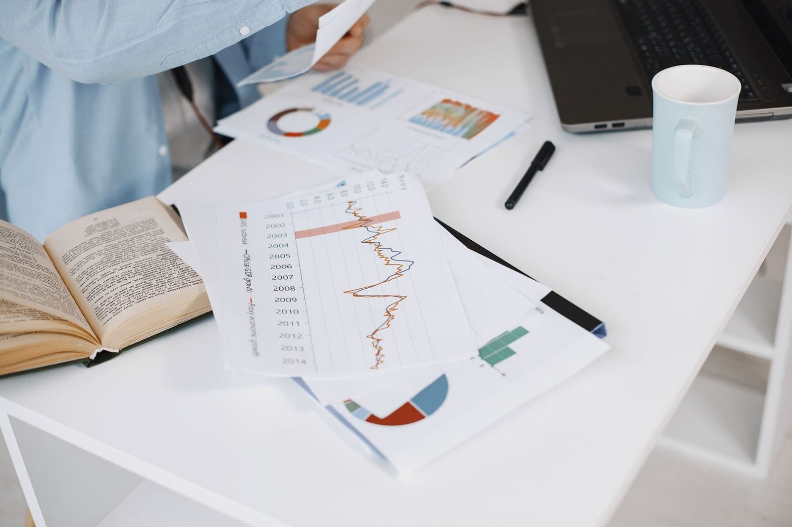Printed charts and a book on a white desk beside a laptop and a mug