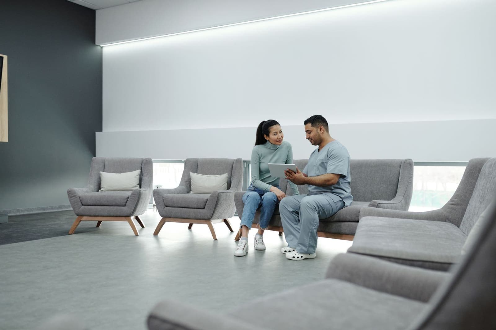 A nurse in scrubs going through a form on a tablet with a patient in a clinic waiting area