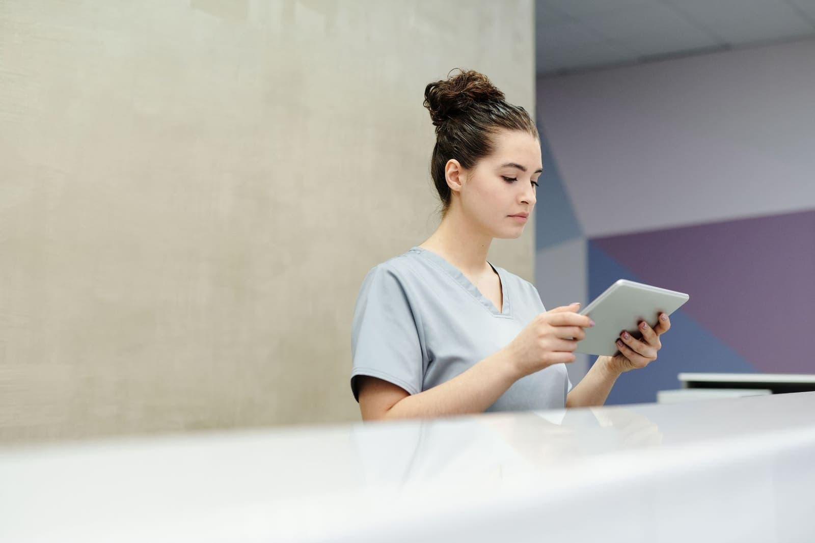 A receptionist in scrubs reading a tablet behind a clinic front desk