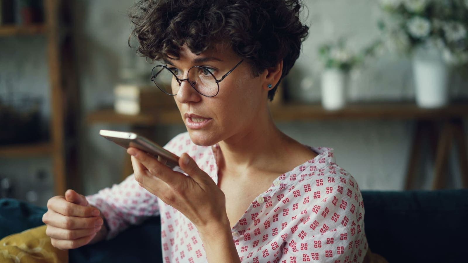 A woman with glasses speaking a question into her phone on a sofa at home