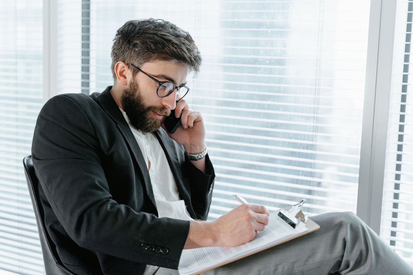 A man on a phone call writing on a clipboard beside an office window