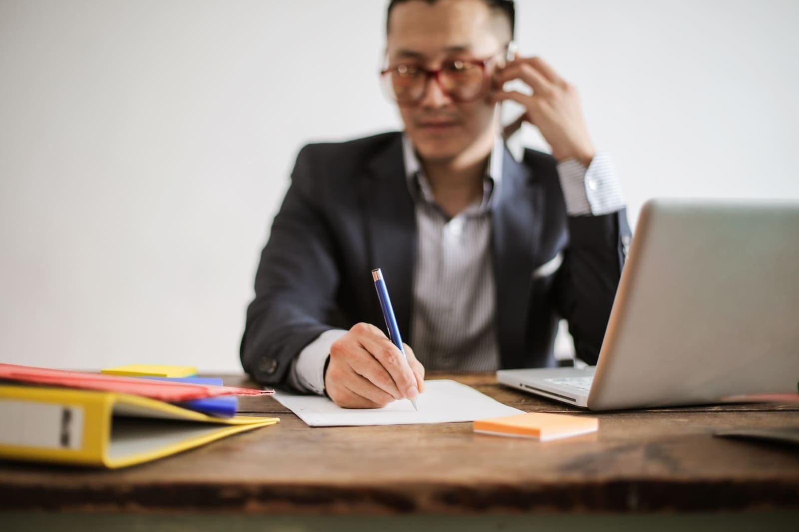 A man on a phone call writing notes beside a laptop at a desk