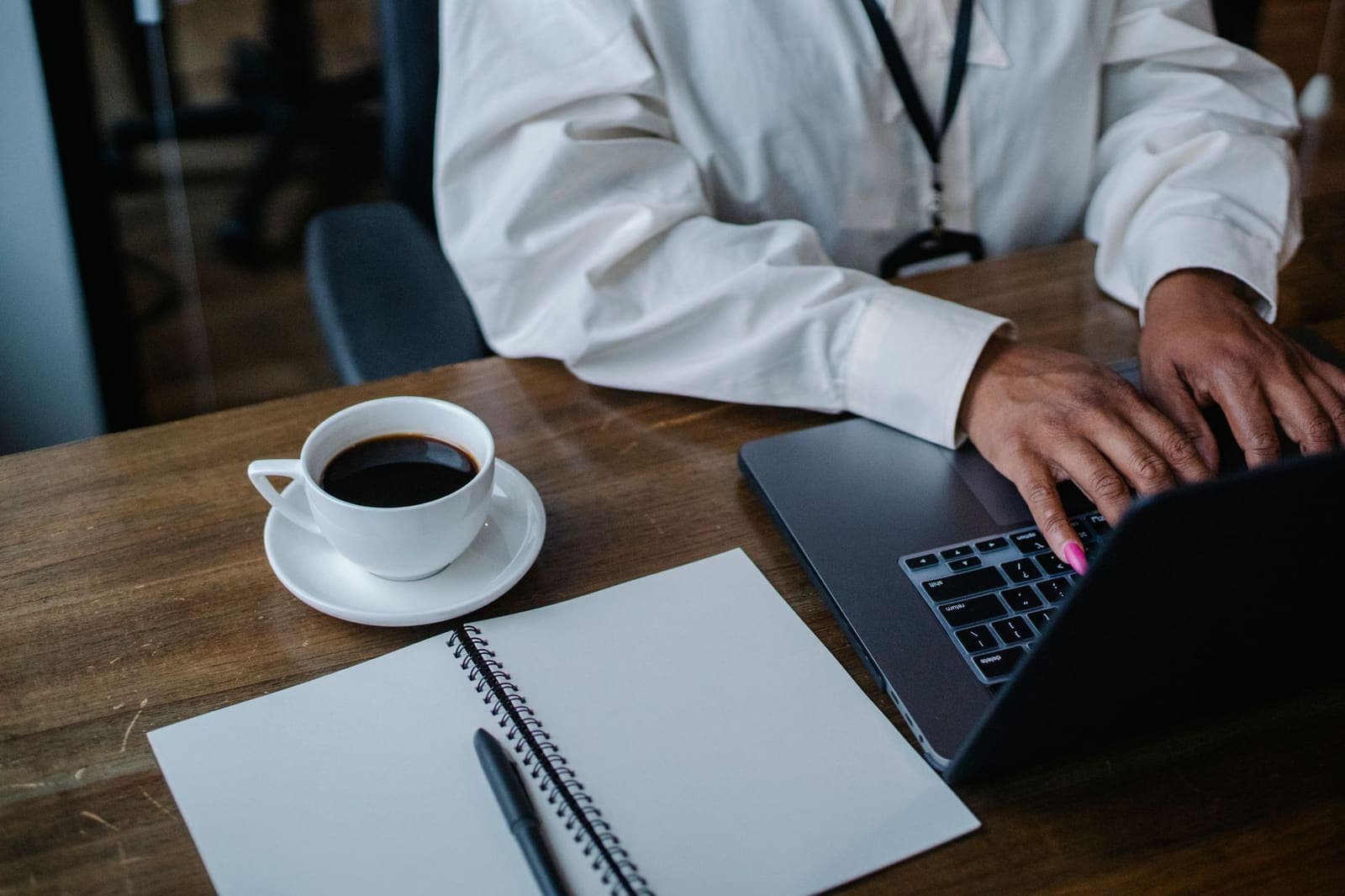 A person in a white shirt typing on a laptop beside a notebook and a cup of coffee