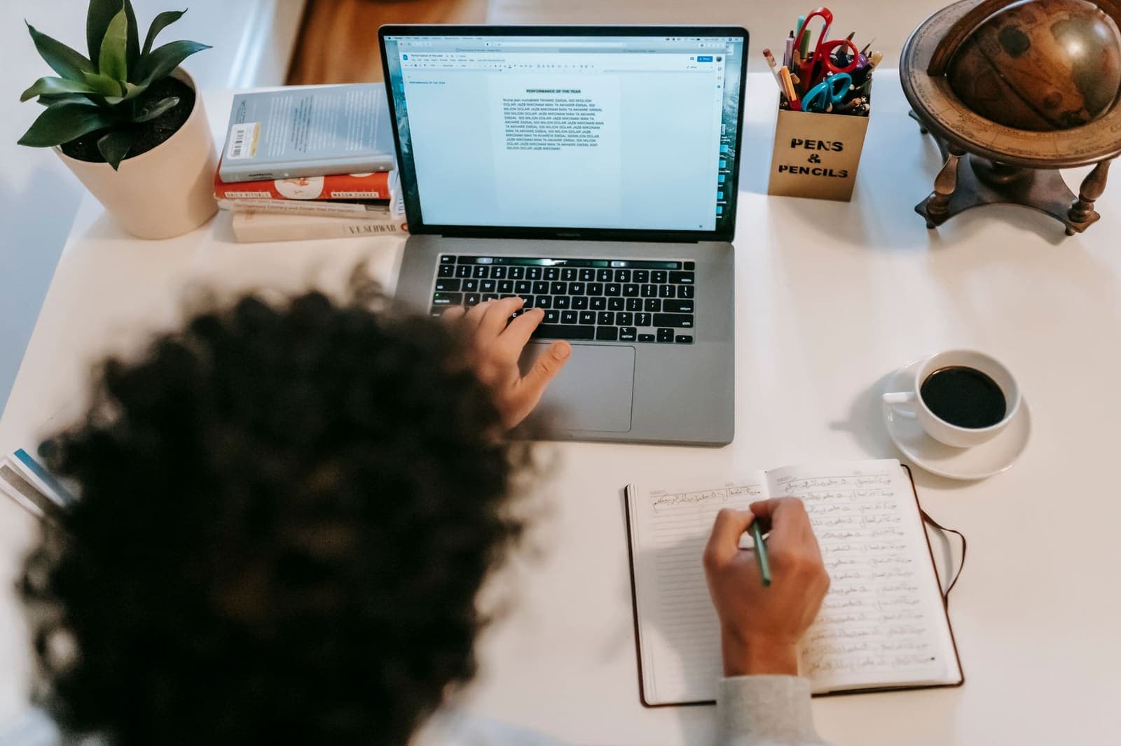 A writer seen from above, typing on a laptop with a notebook and a coffee beside it