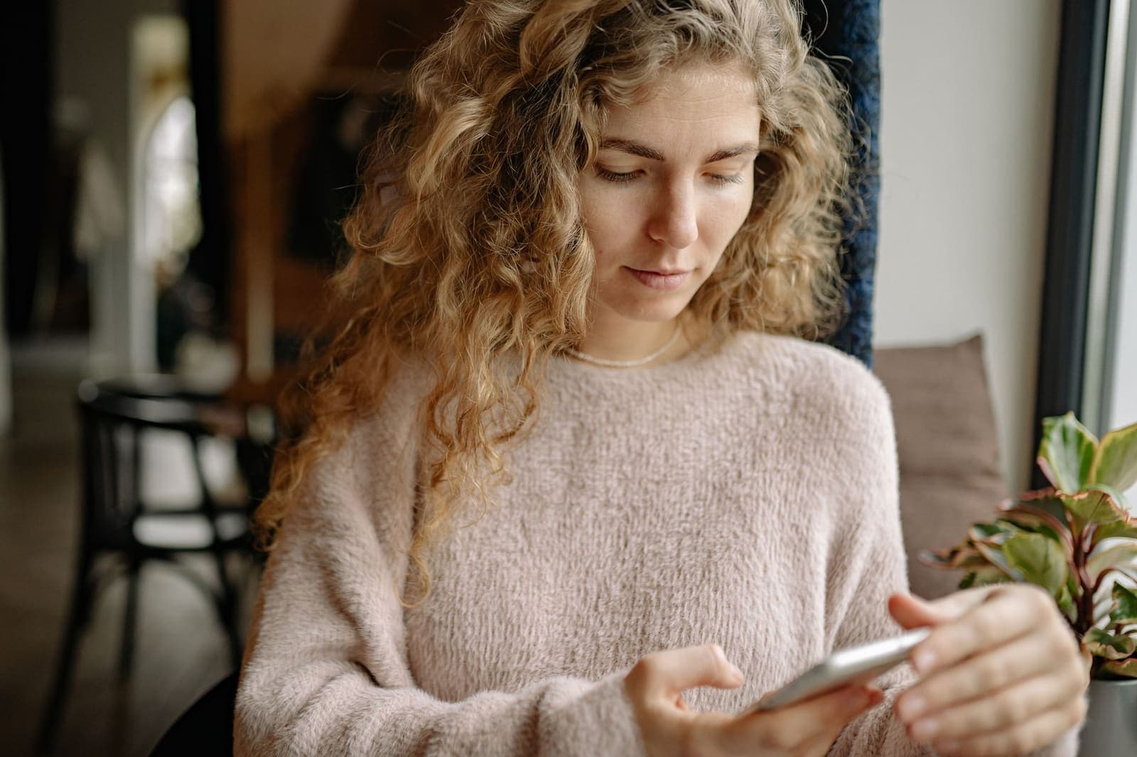 A woman in a soft sweater reading a message on her phone by a window