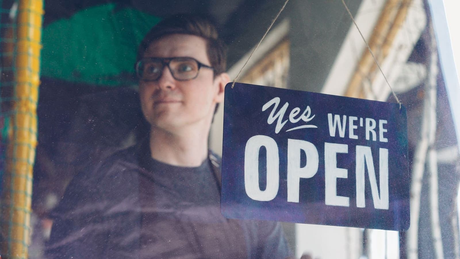A shop owner behind a glass door with a hand-lettered sign reading Yes, We're Open