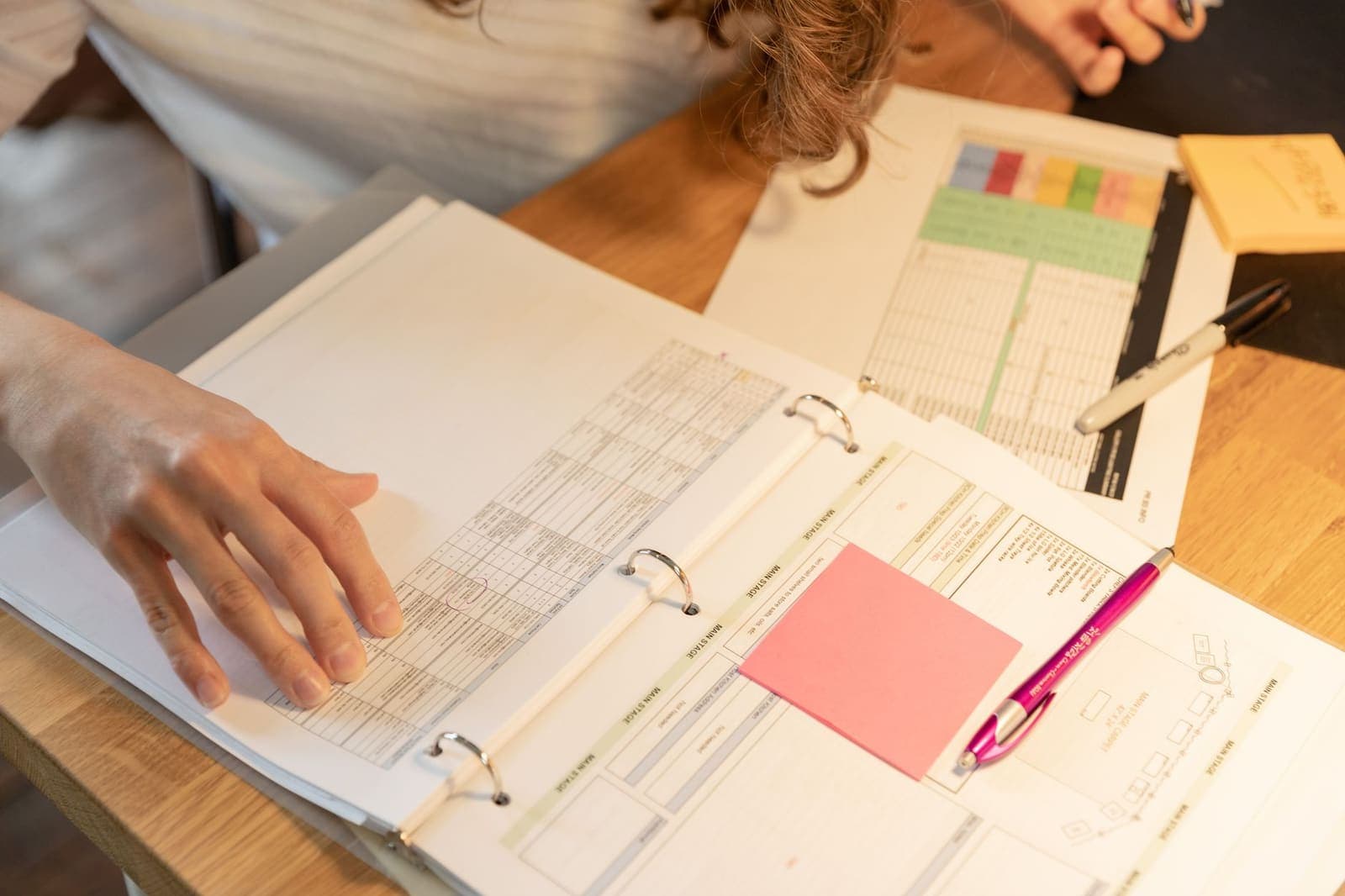 An open binder of schedules and forms on a desk with sticky notes and a pen