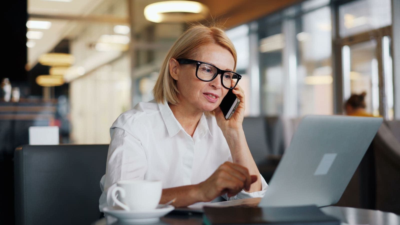A woman in glasses on a phone call at a cafe table with her laptop open