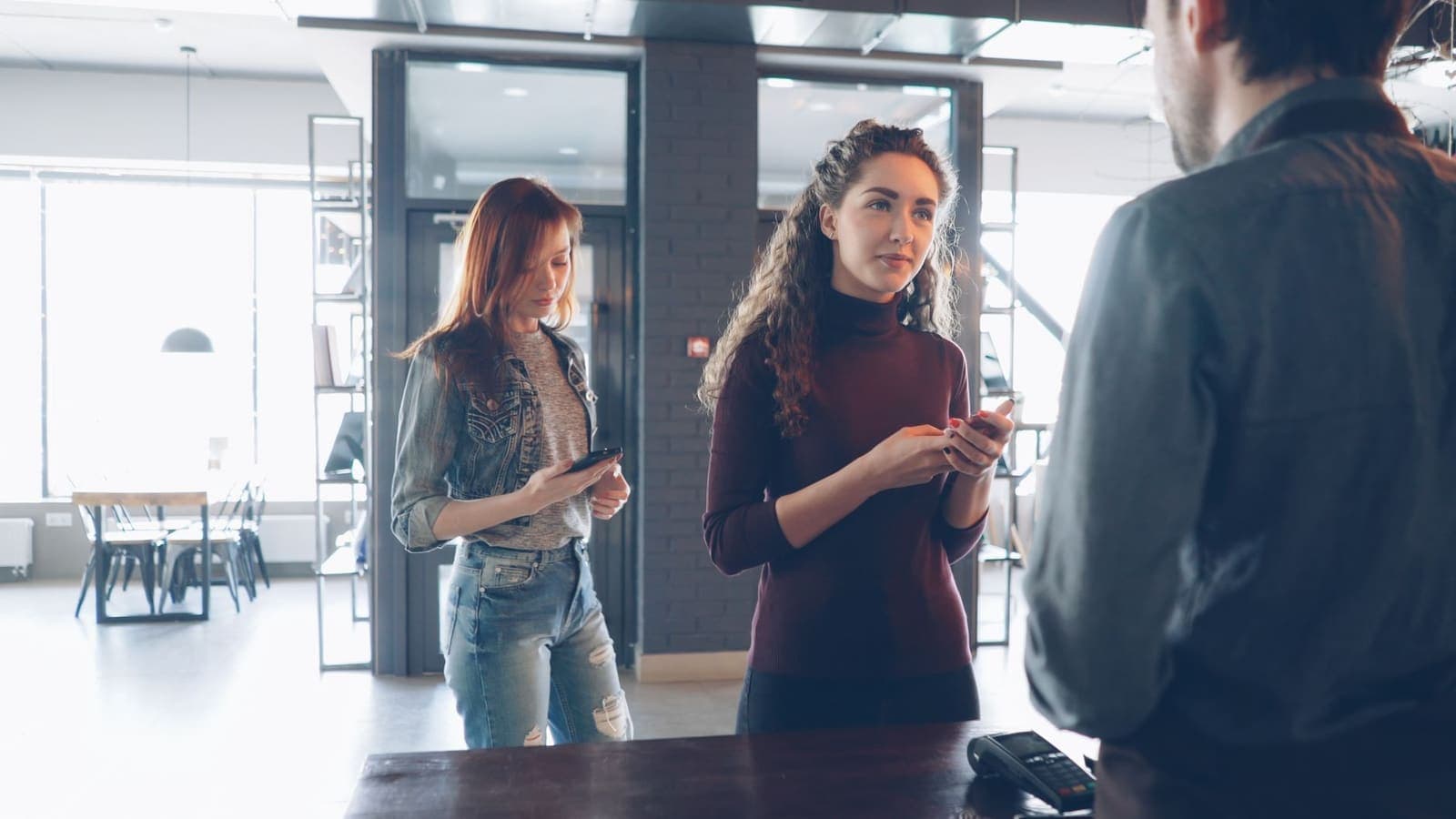 Two customers at a cafe counter with their phones out while the owner takes their order