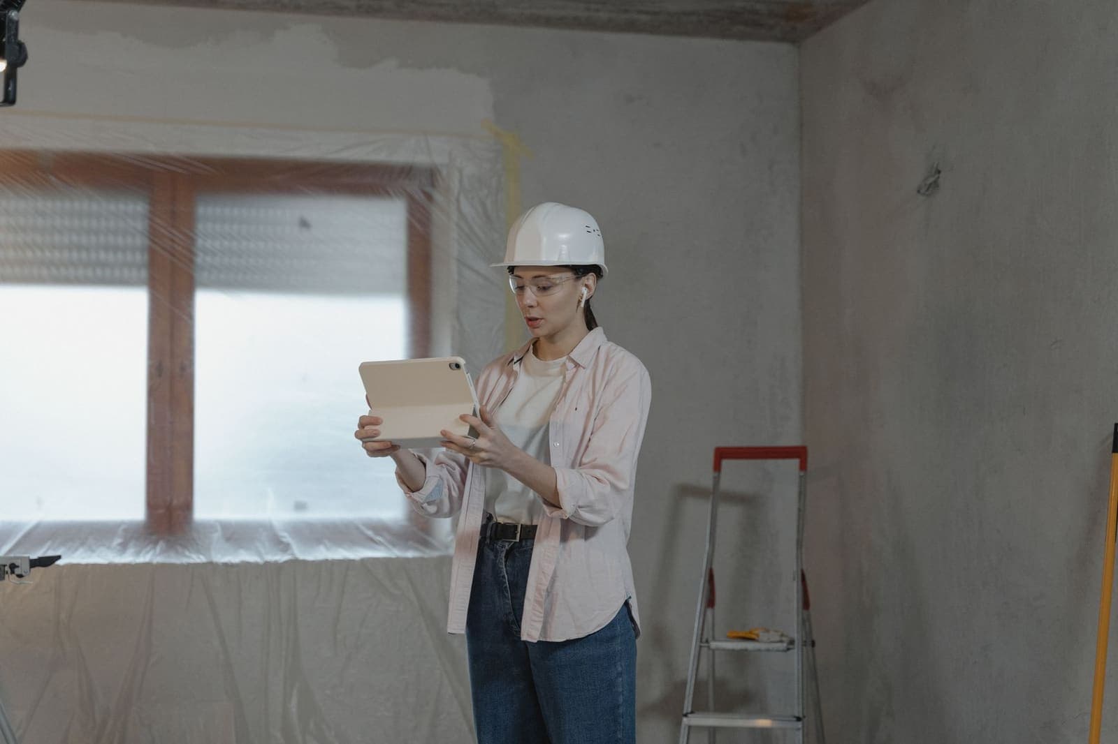 A woman in a hard hat reading a tablet in an unfinished room