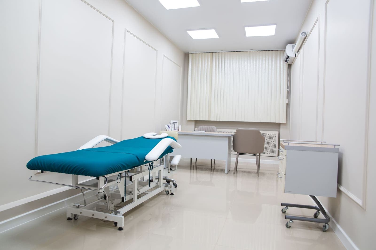 An empty, clean examination room with a teal treatment table, a desk with two chairs and a rolling cabinet