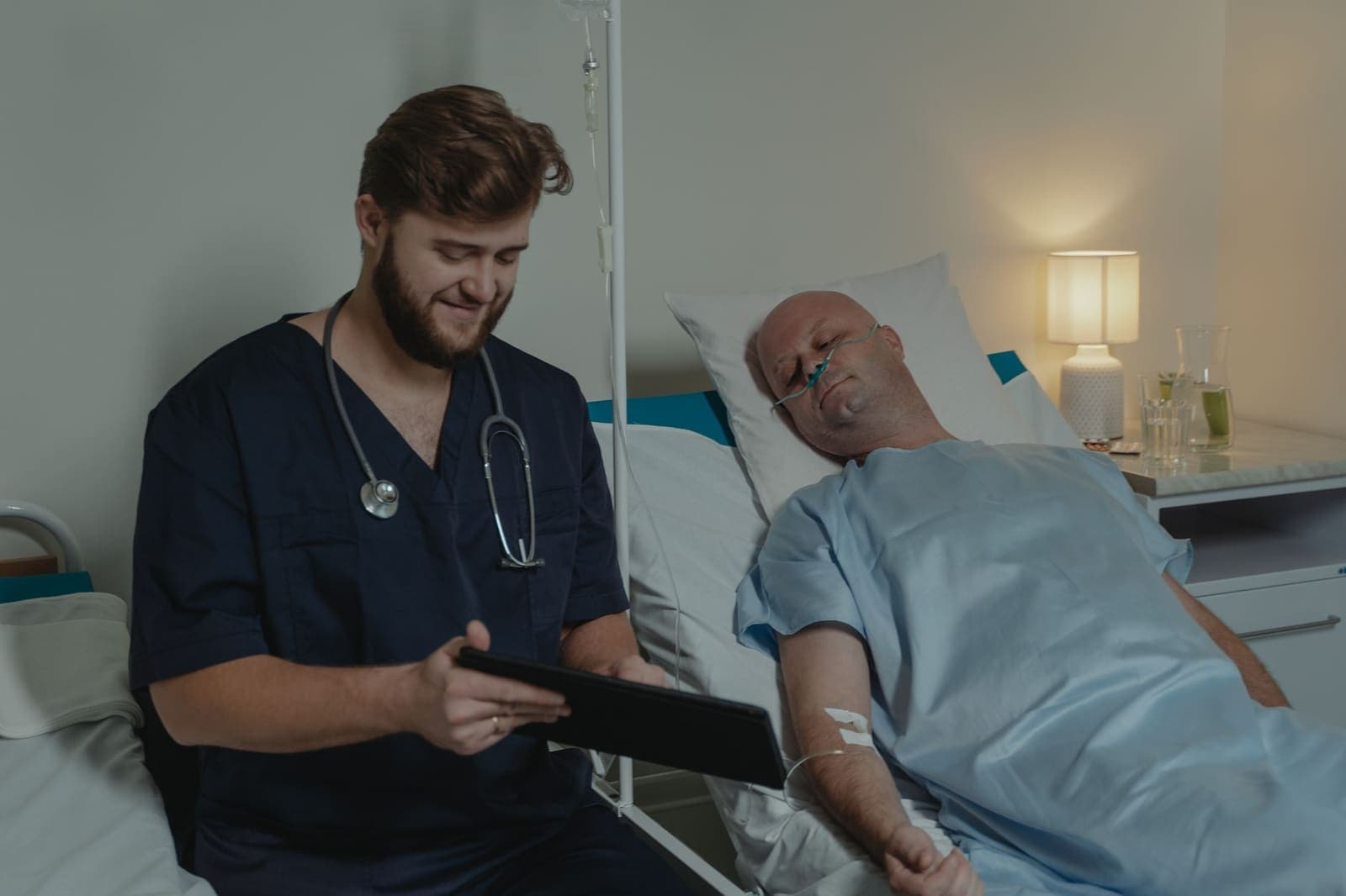 A clinician in navy scrubs reviewing a tablet while sitting beside a patient in a hospital bed
