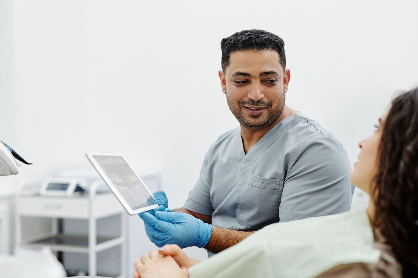 A clinician in grey scrubs and blue gloves showing a tablet to a patient seated in an exam chair