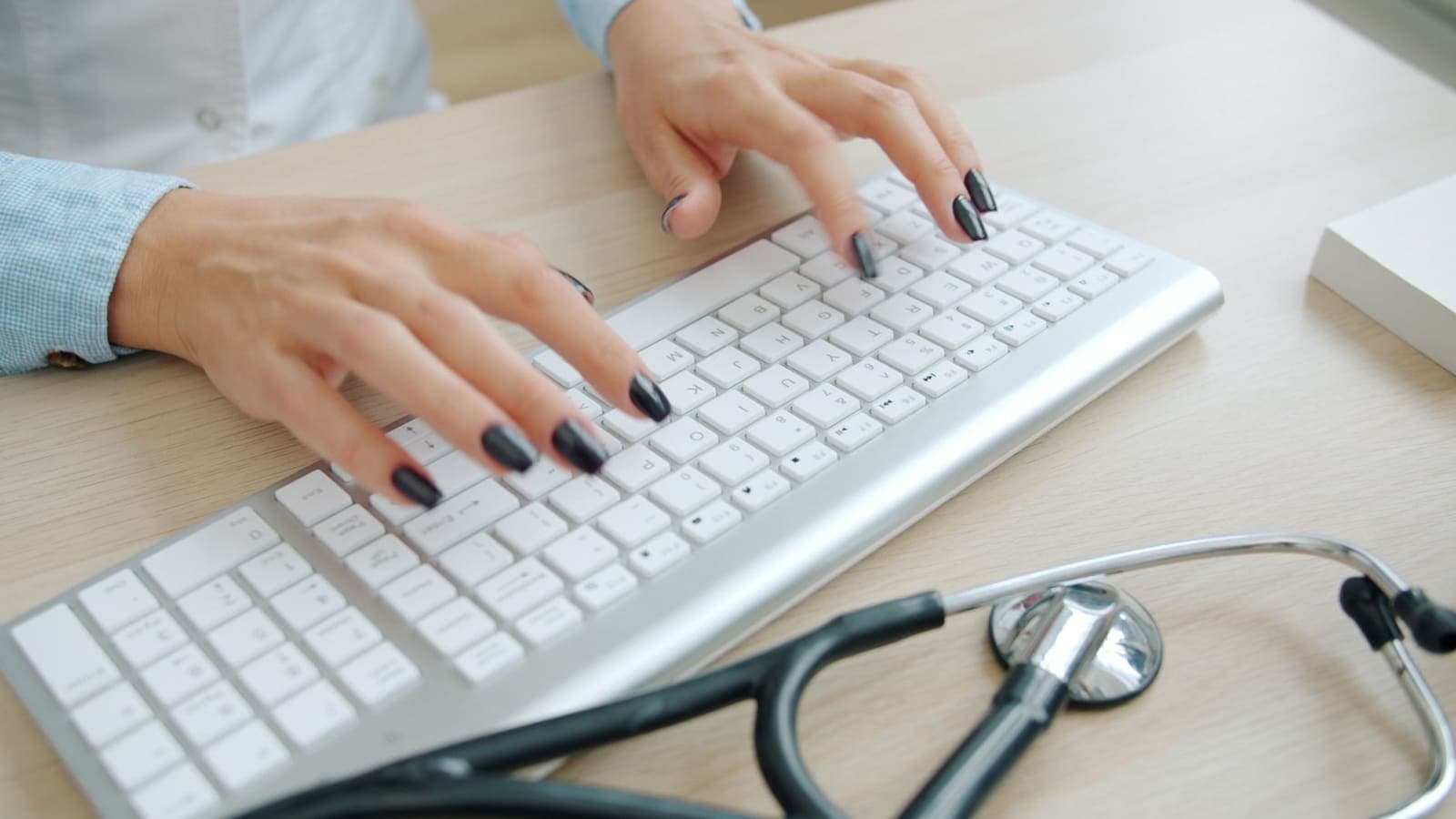 Close-up of hands typing on a white keyboard on a wooden desk beside a stethoscope