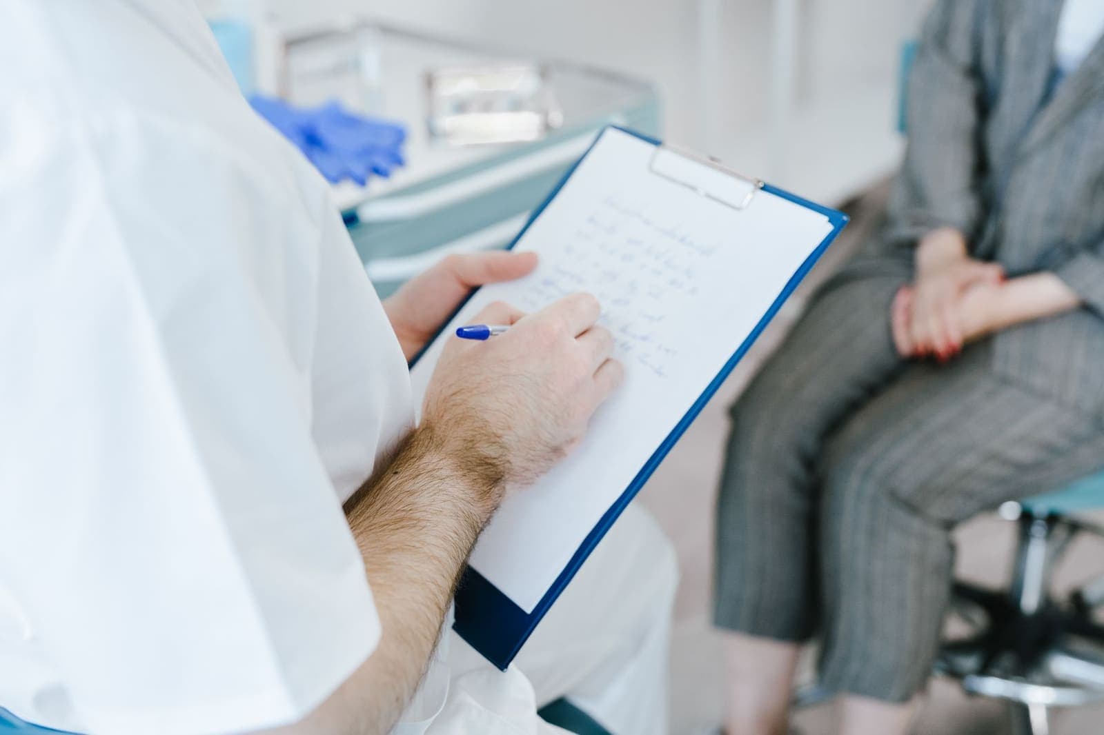 A clinician in white writing notes on a blue clipboard while a seated person waits out of focus