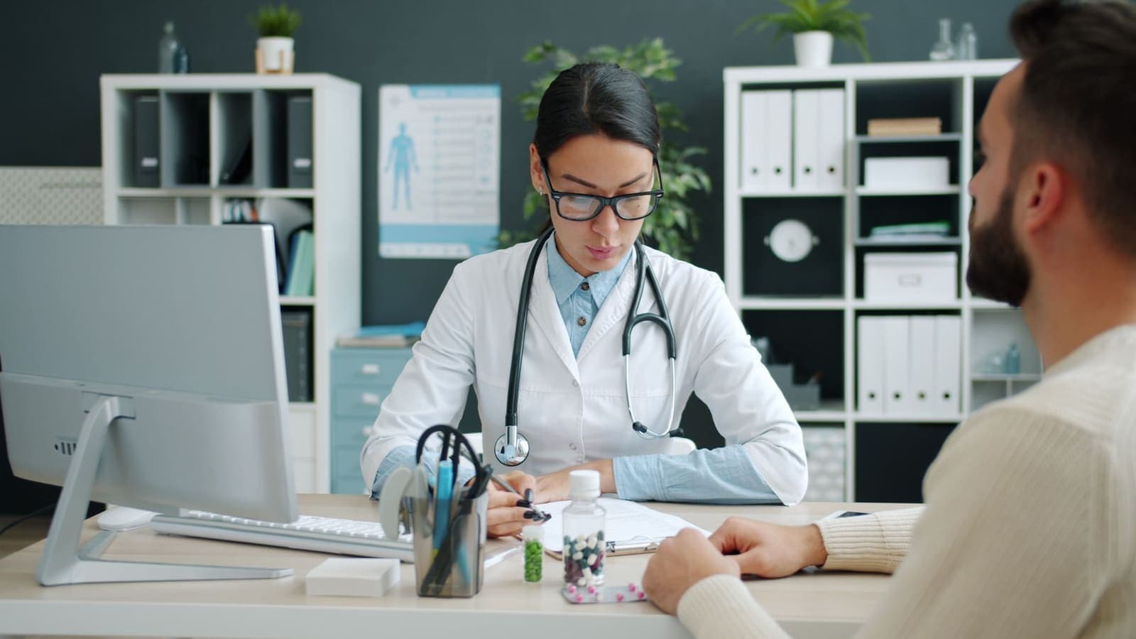 A doctor in a white coat and glasses writing notes across a desk from a patient, with medication bottles on the desk